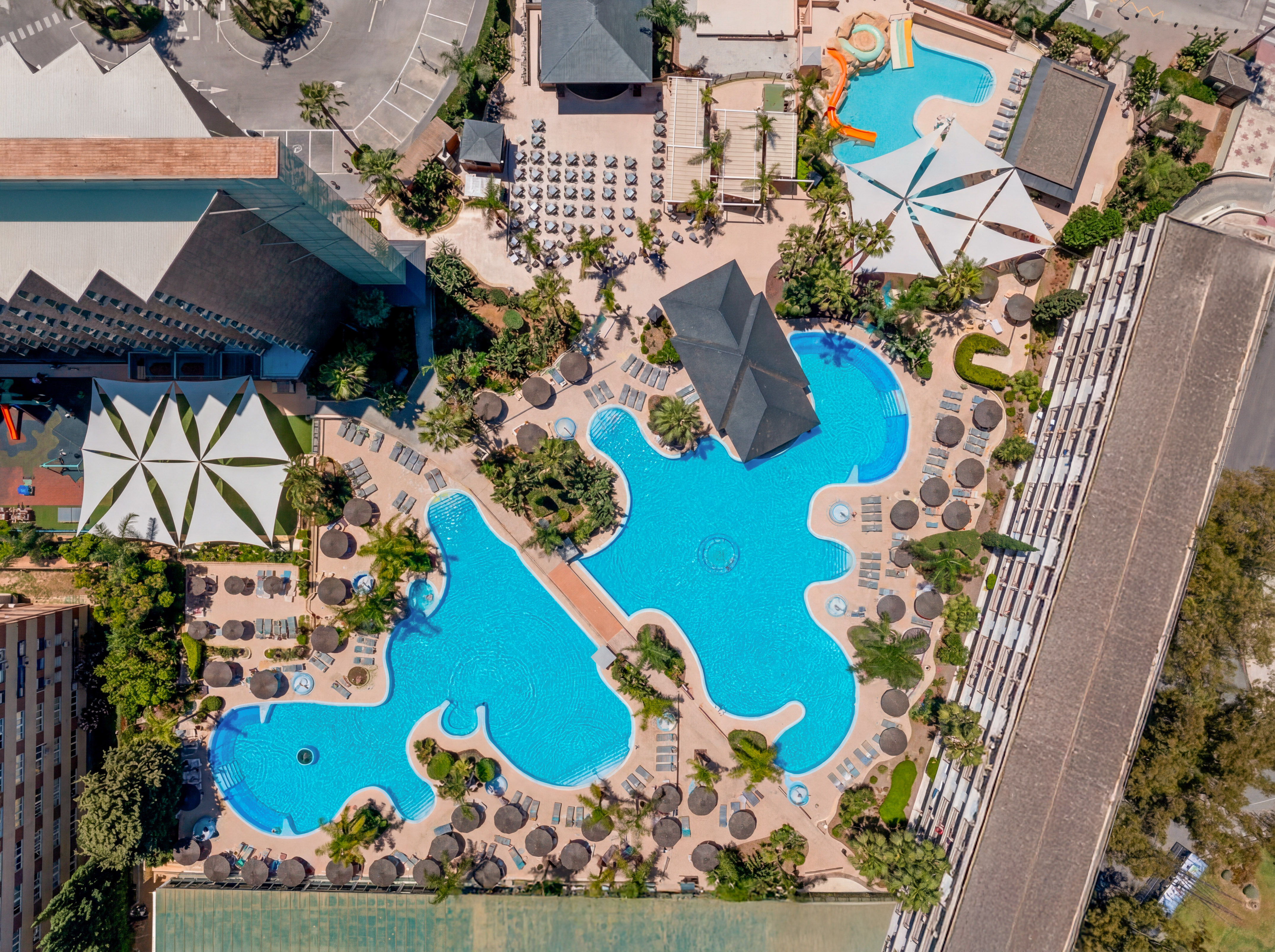 Resort aerial: interconnected swimming pools, sun loungers, palm trees, water slides.
