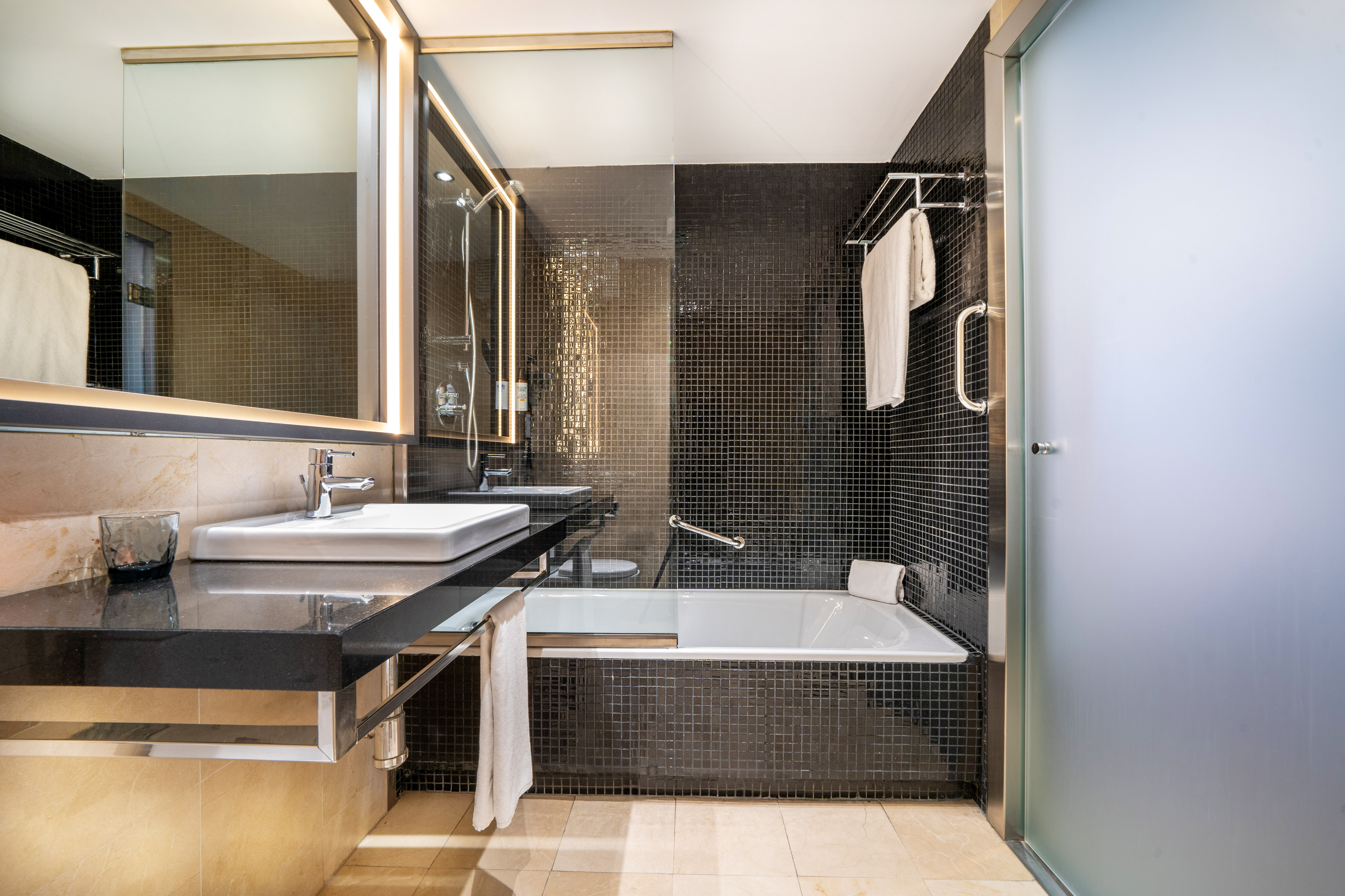 Modern hotel bathroom with black mosaic tiles, bathtub, vanity, and mirror.