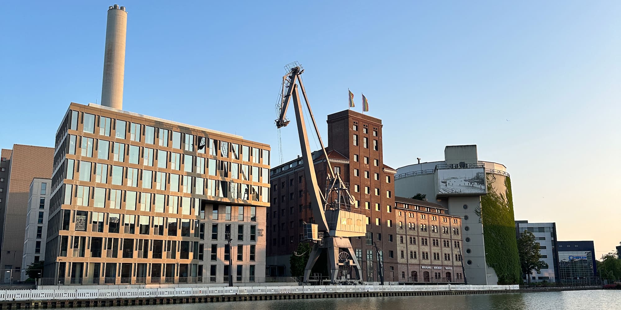 Waterfront buildings, industrial crane, clear sky, with reflections on water.