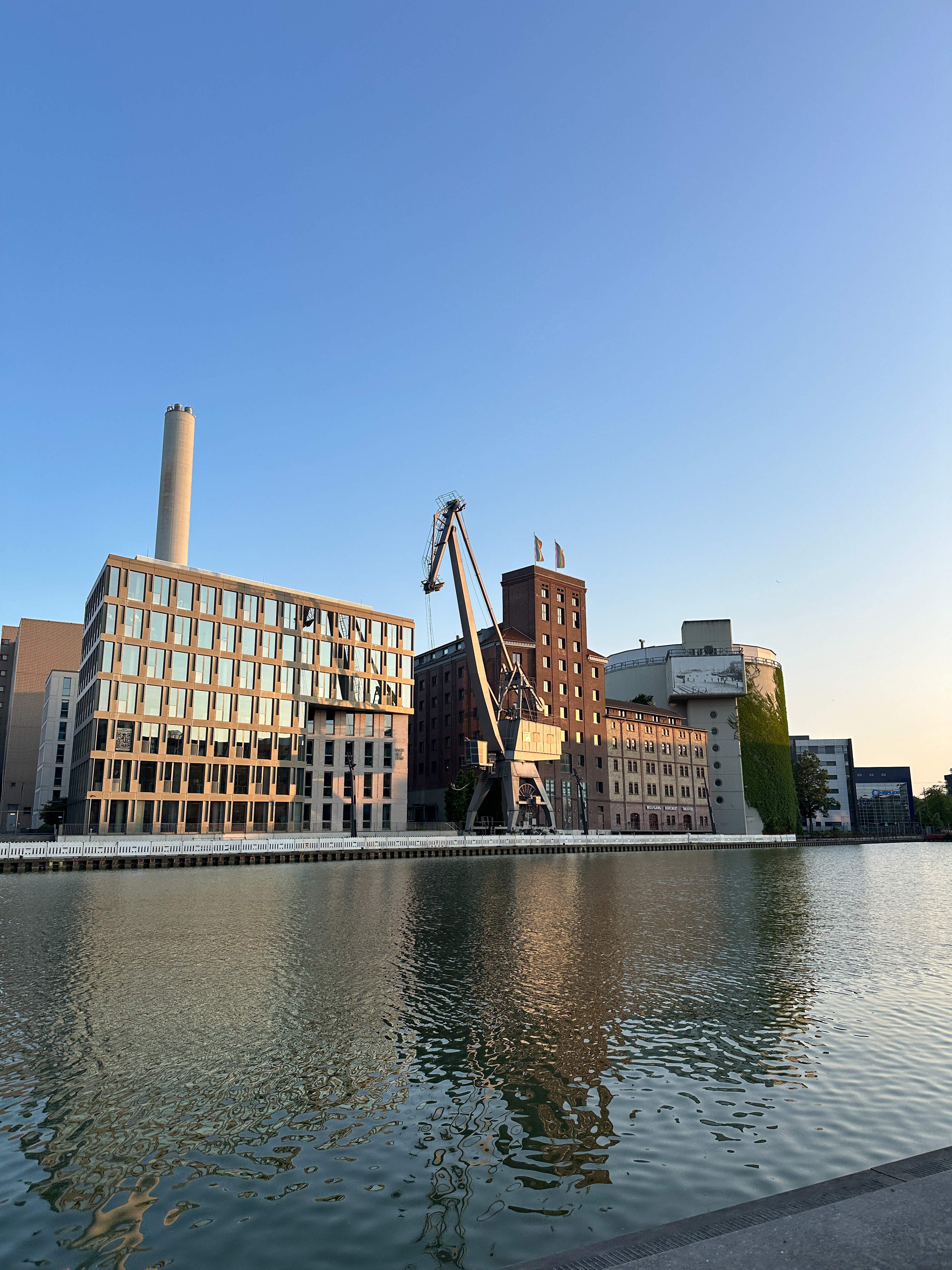 Waterfront buildings, industrial crane, clear sky, with reflections on water.