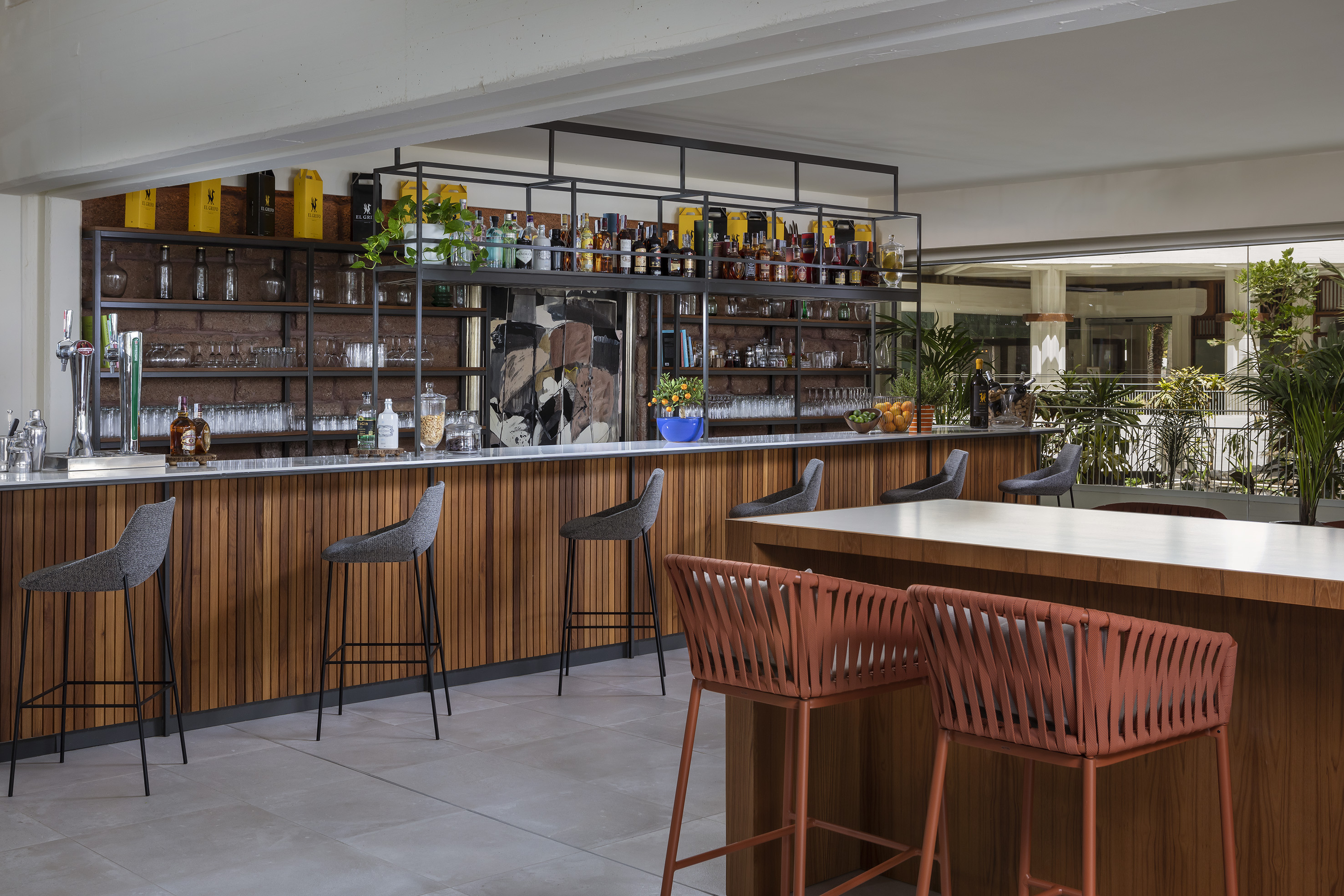 Modern hotel bar with wooden counter, bar stools, liquor bottles, and plants.