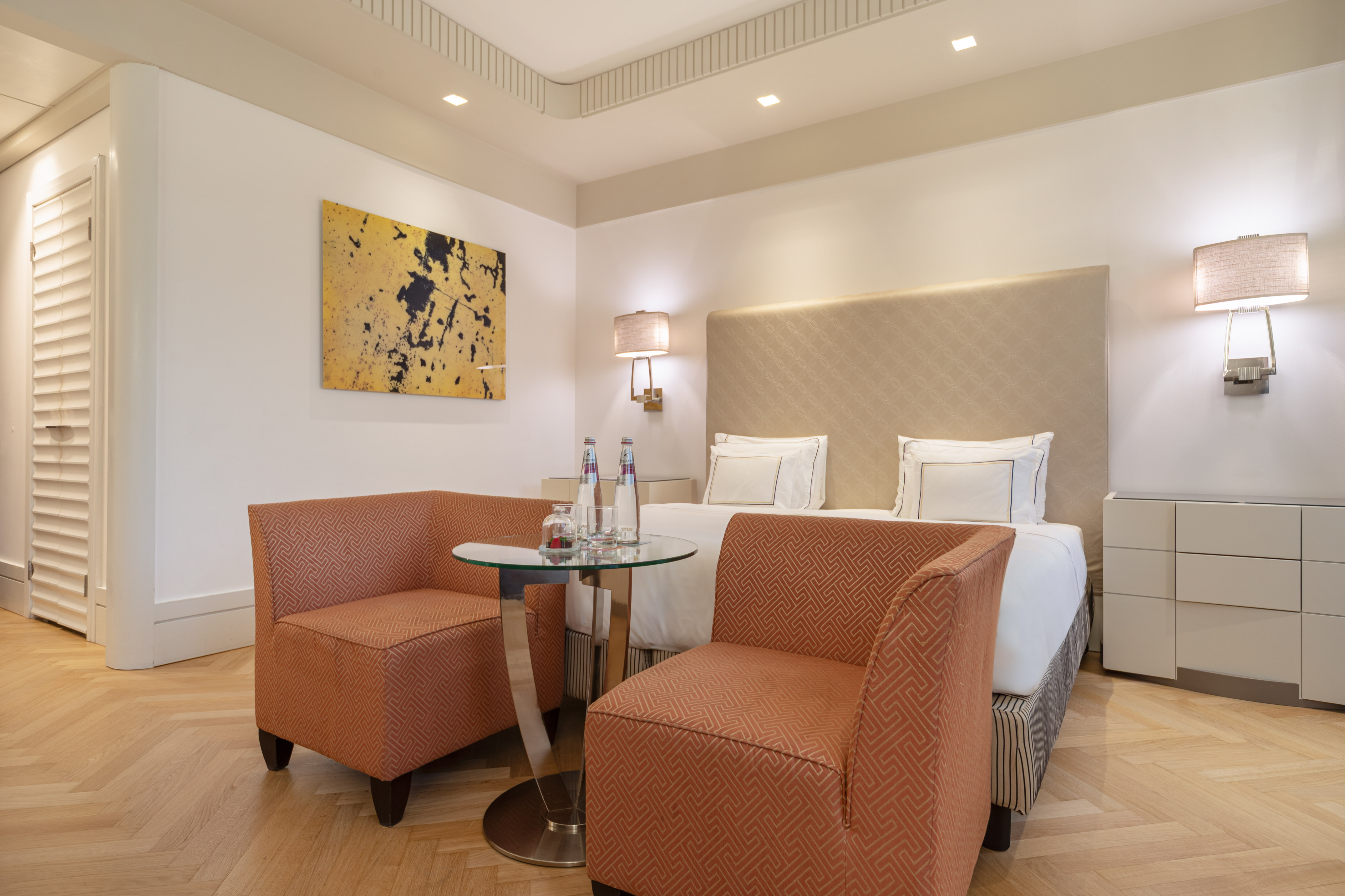 Hotel bedroom with king bed, two orange armchairs, and contemporary art.