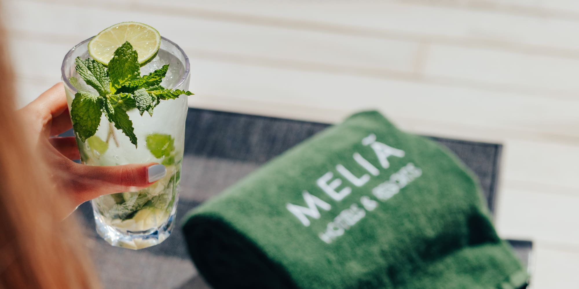 Hand holding a refreshing mojito by Melia Hotels resort pool, green towel.