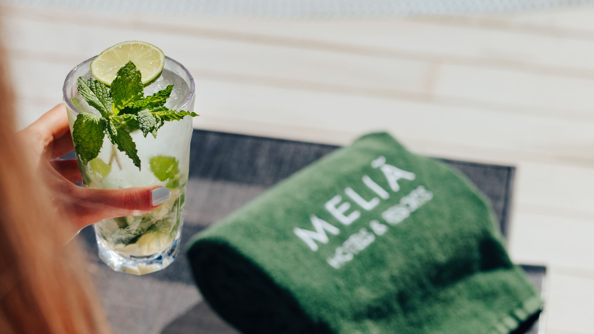 Hand holding a refreshing mojito by Melia Hotels resort pool, green towel.
