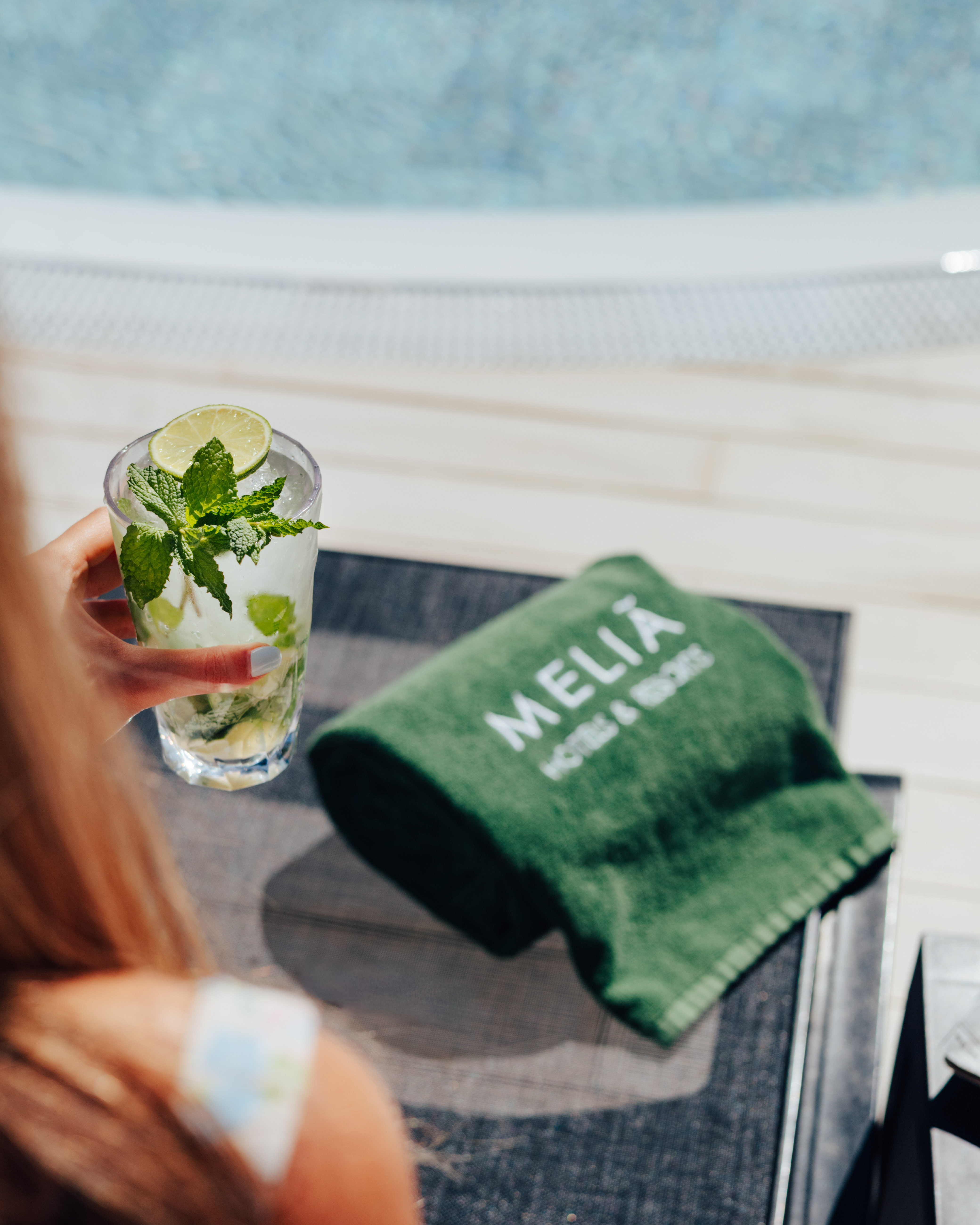 Hand holding a refreshing mojito by Melia Hotels resort pool, green towel.