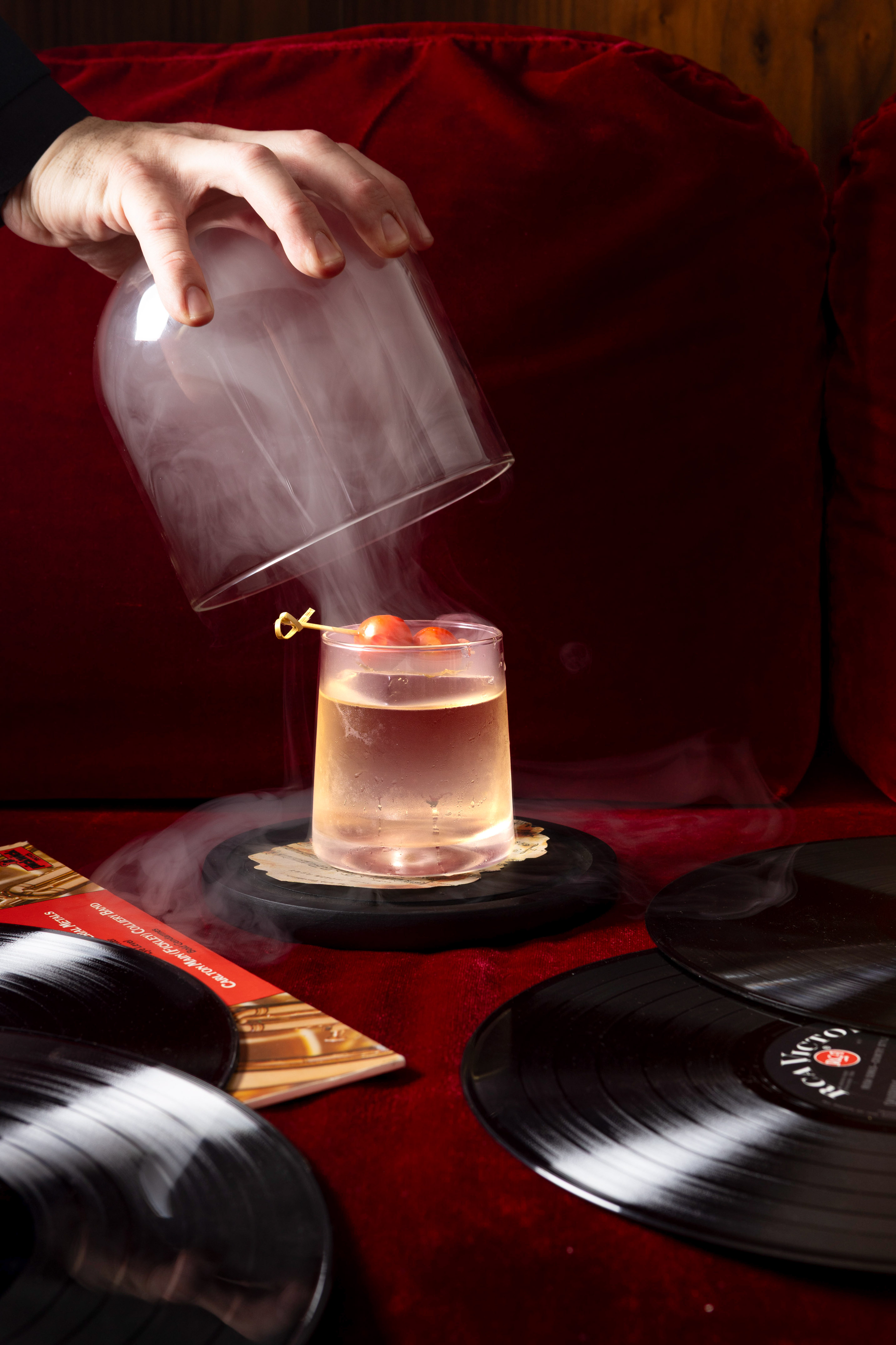 Hand removes smoky cloche from cherry cocktail, vinyl records on red velvet.