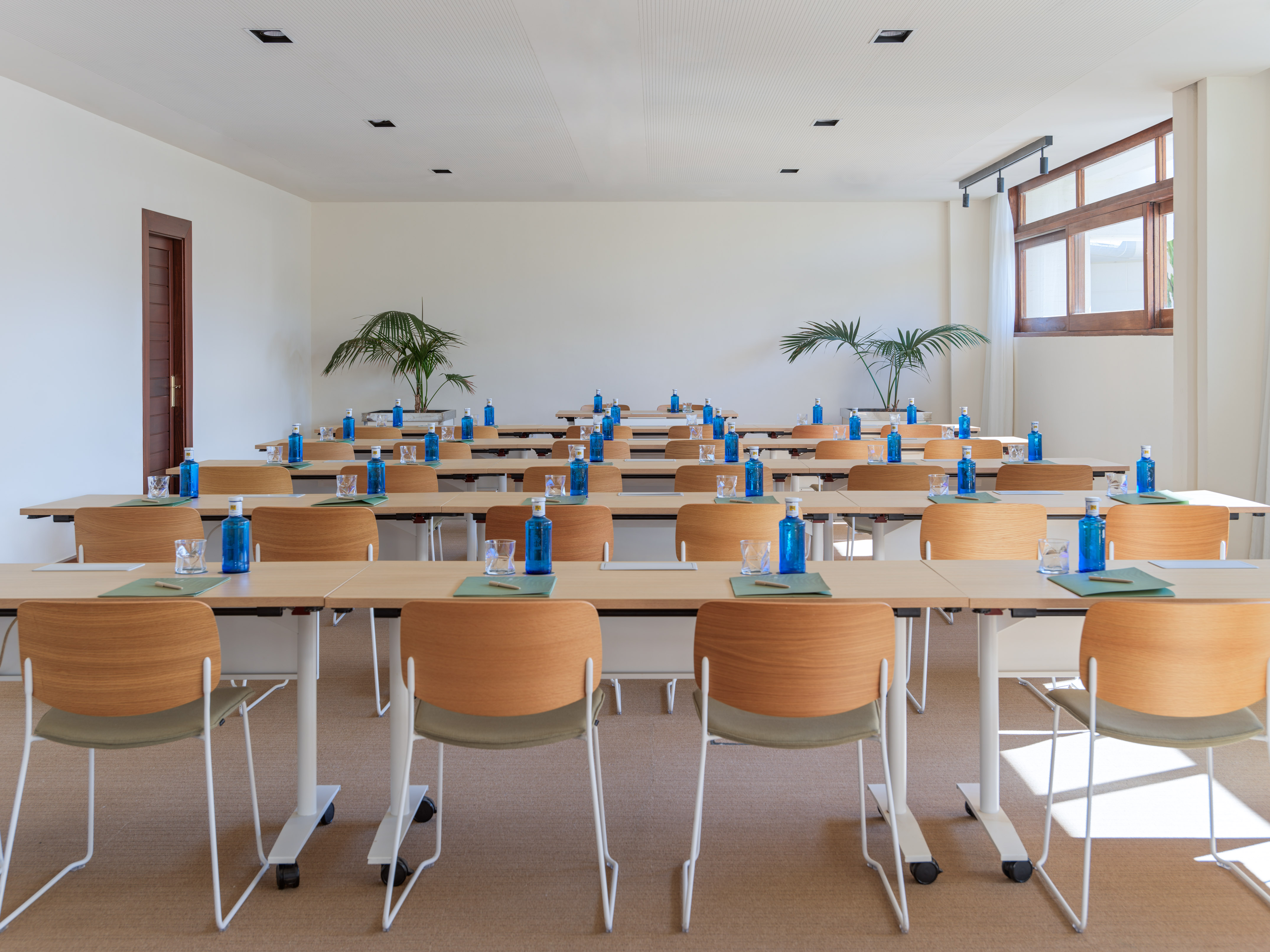 Modern conference room with rows of tables, chairs, blue water bottles, and plants.