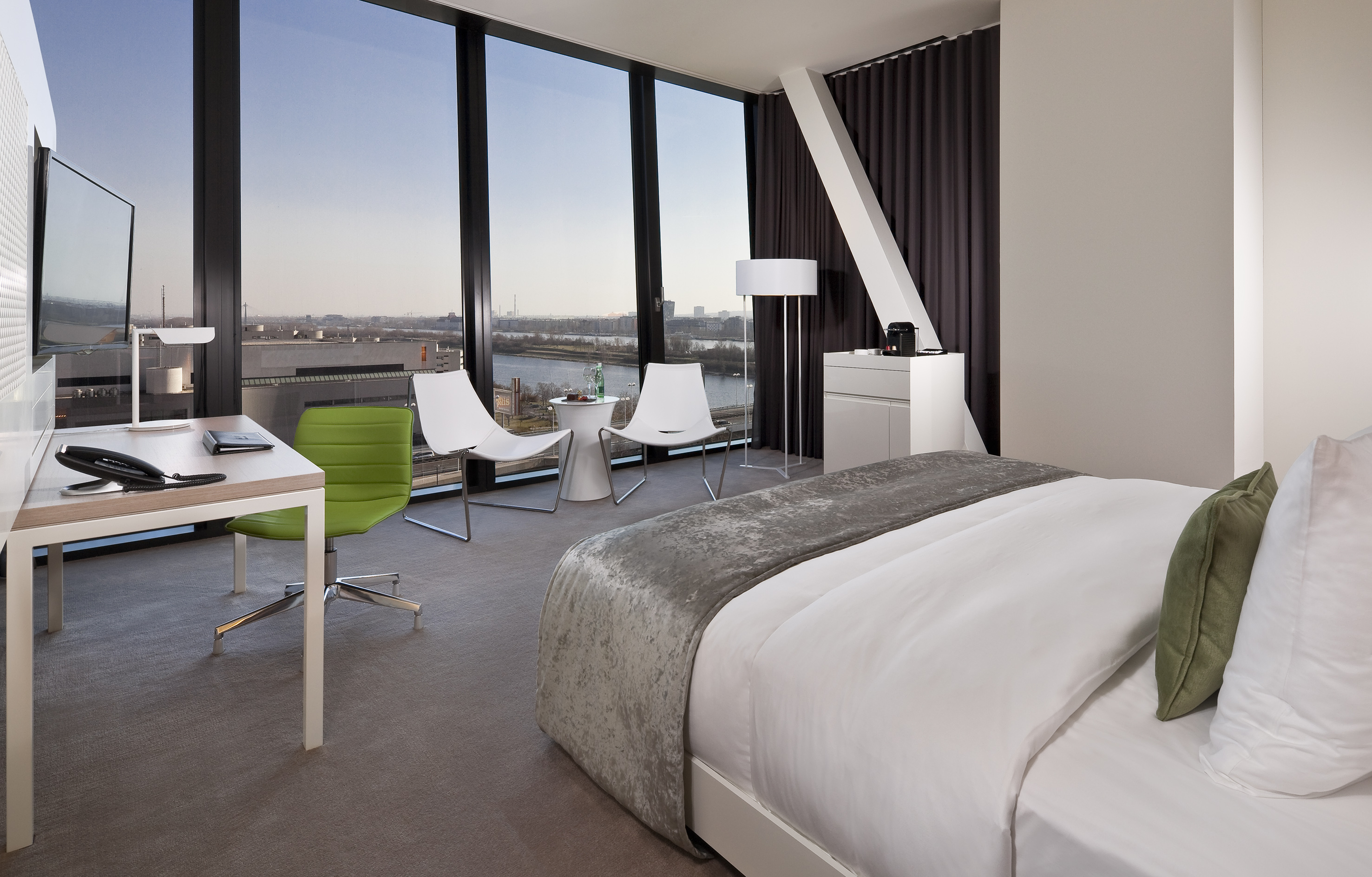 Modern hotel room with river view, bed, desk, and lounge chairs by the window.