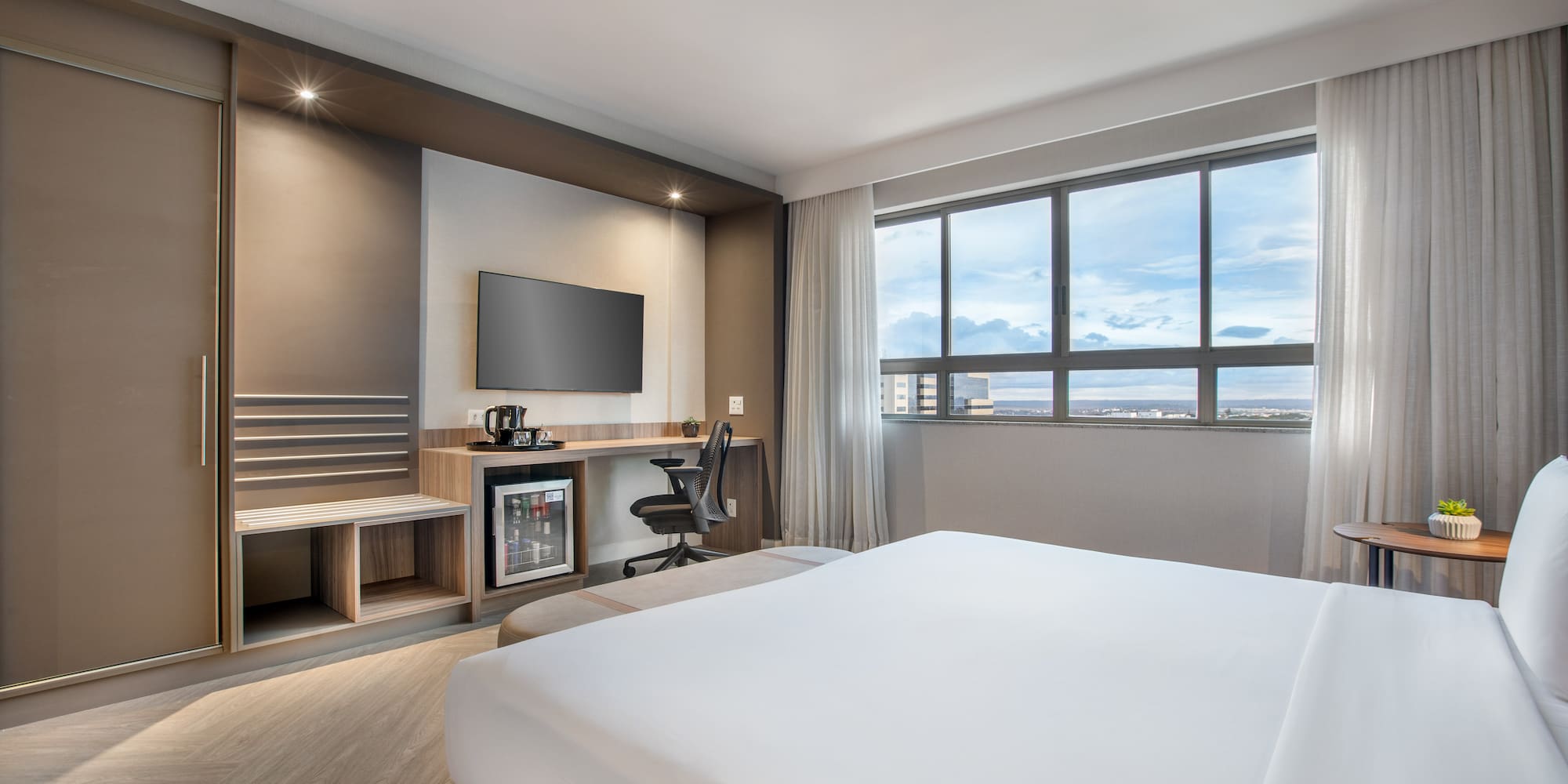 Modern hotel room with white bed, TV, desk, minibar, and window city view.