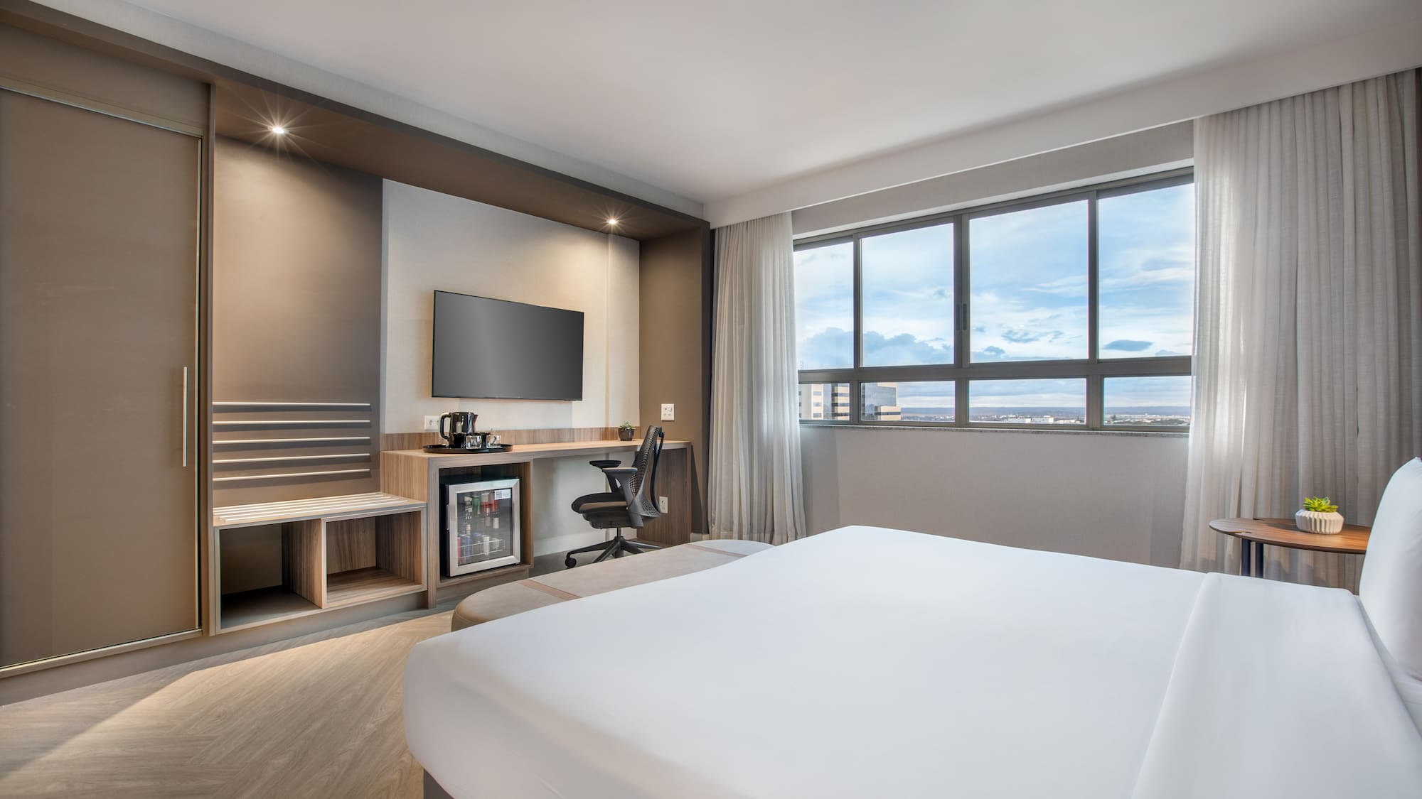Modern hotel room with white bed, TV, desk, minibar, and window city view.