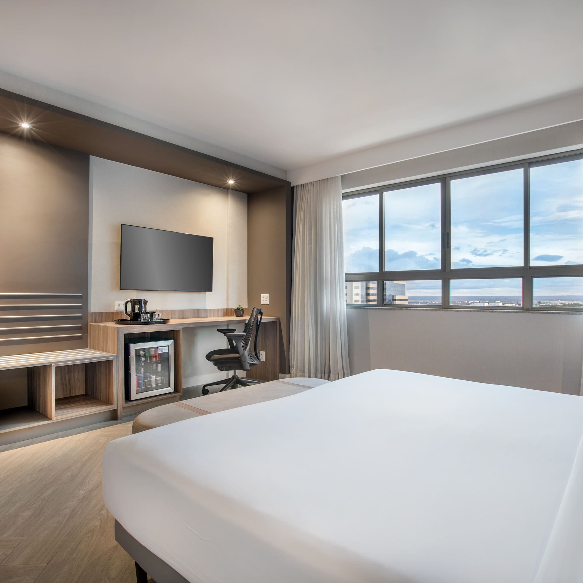 Modern hotel room with white bed, TV, desk, minibar, and window city view.