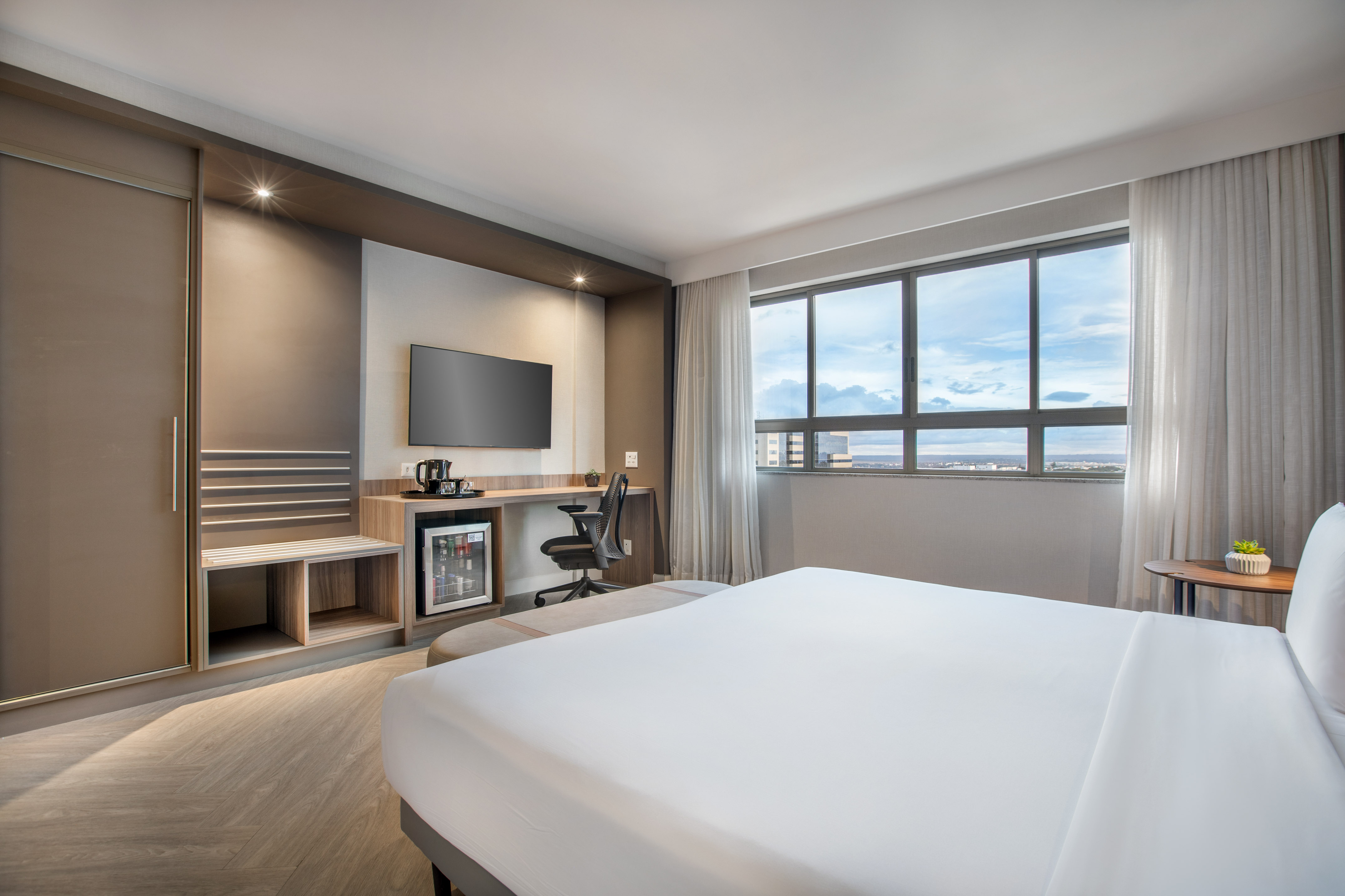 Modern hotel room with white bed, TV, desk, minibar, and window city view.