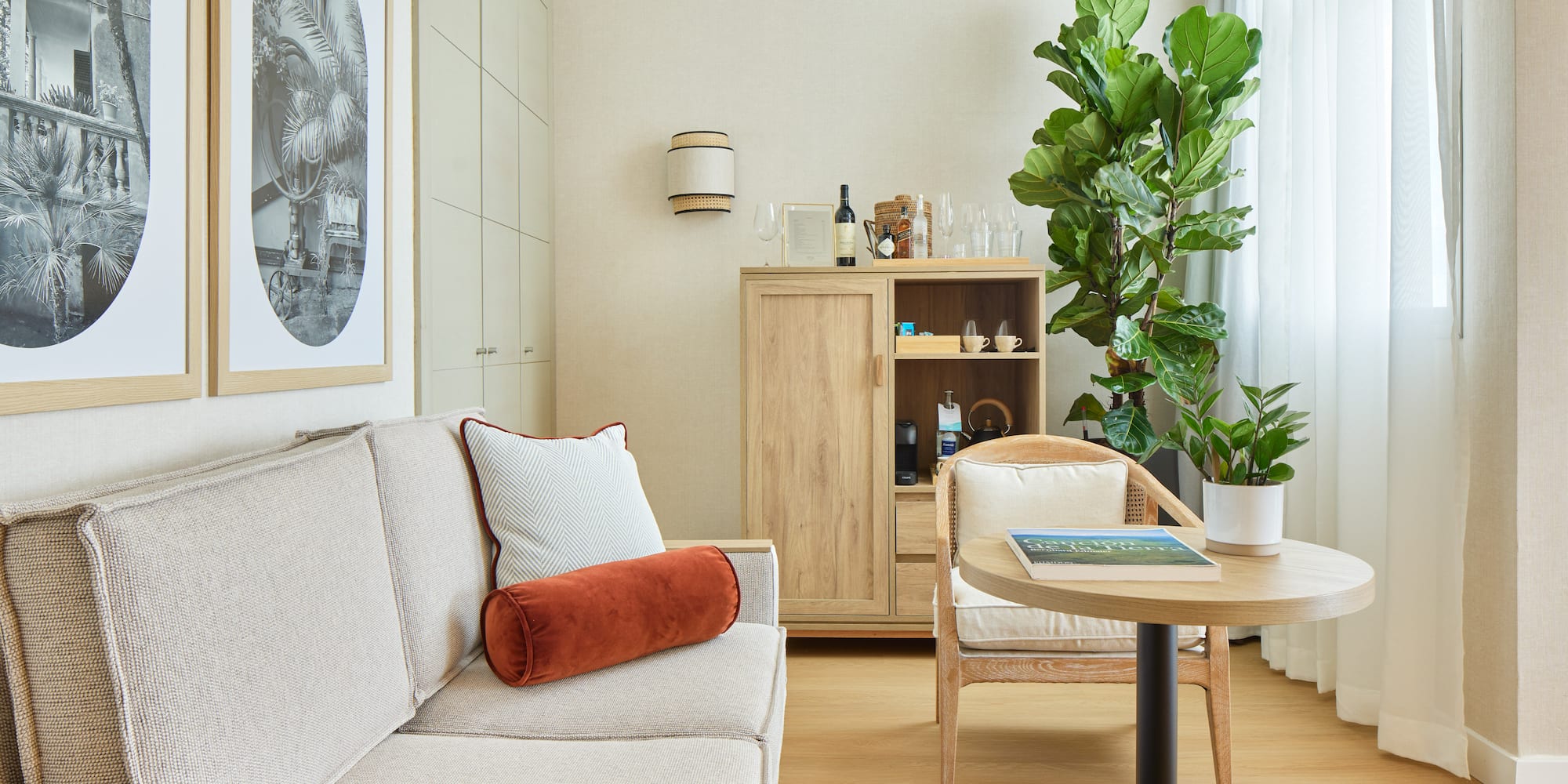 Hotel guest room with neutral sofa, wooden cabinet, and fiddle leaf fig plant.