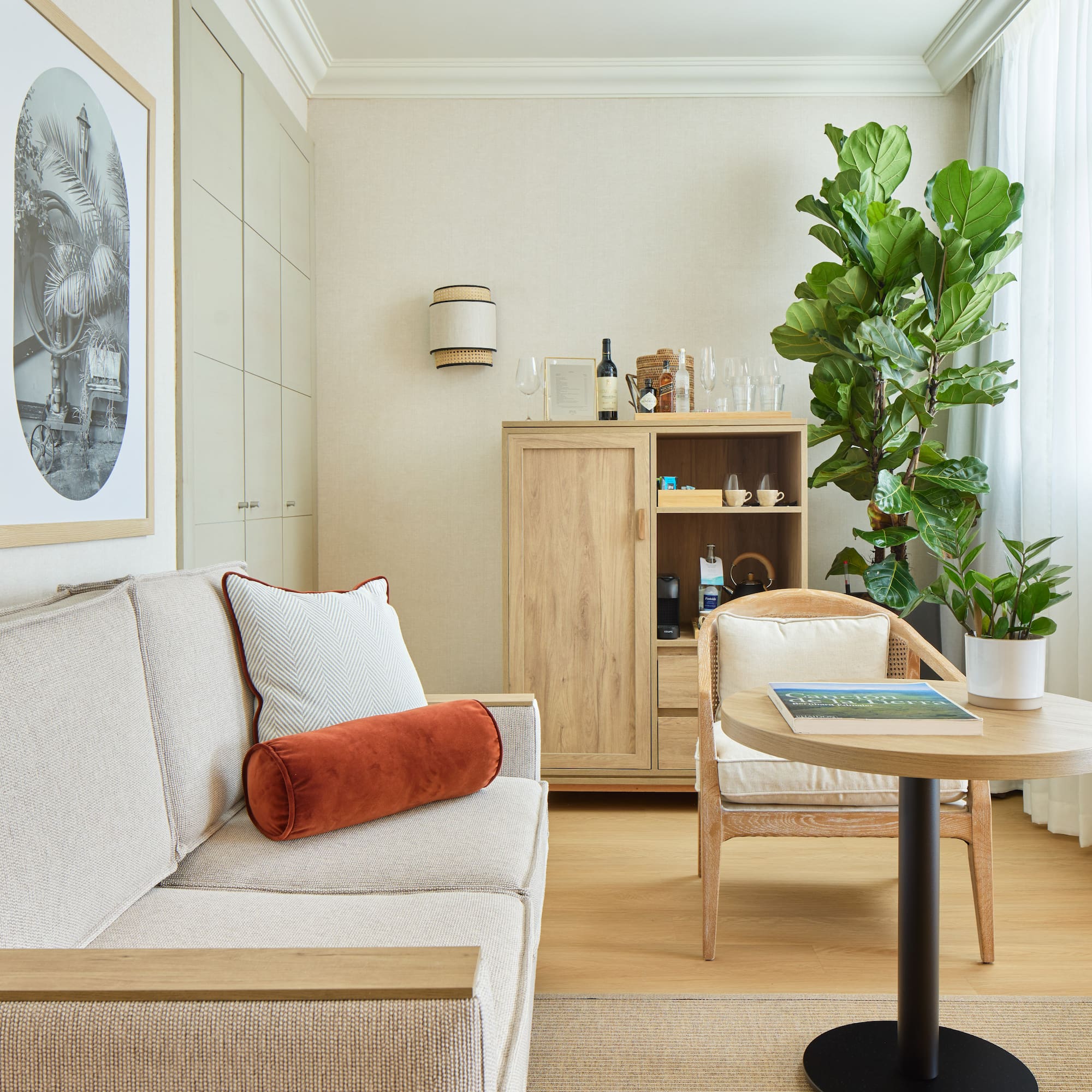 Neutral hotel suite with sofa, wooden cabinet, plants, and framed art.