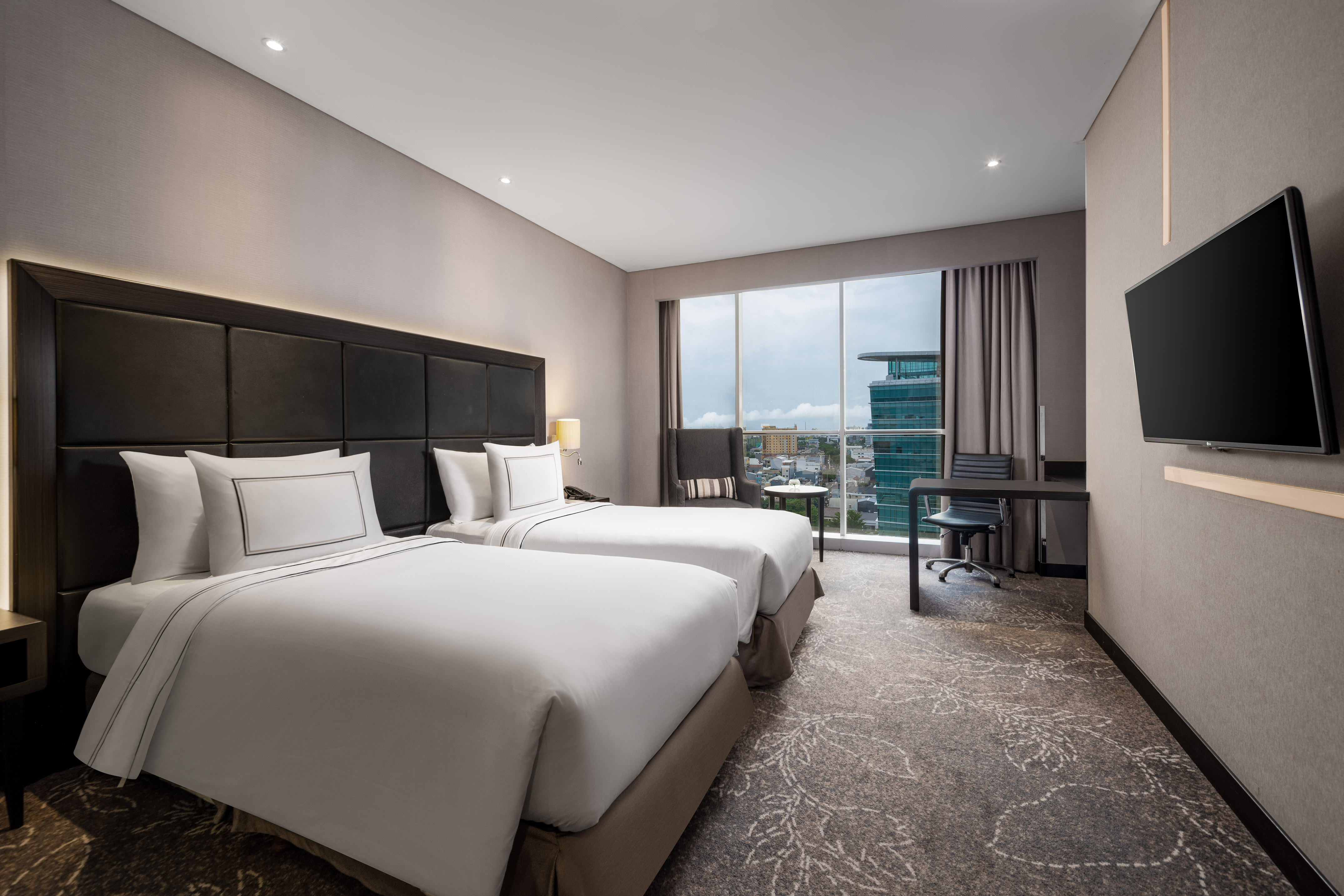 Bright modern hotel room with two twin beds, city skyline view, and workspace.
