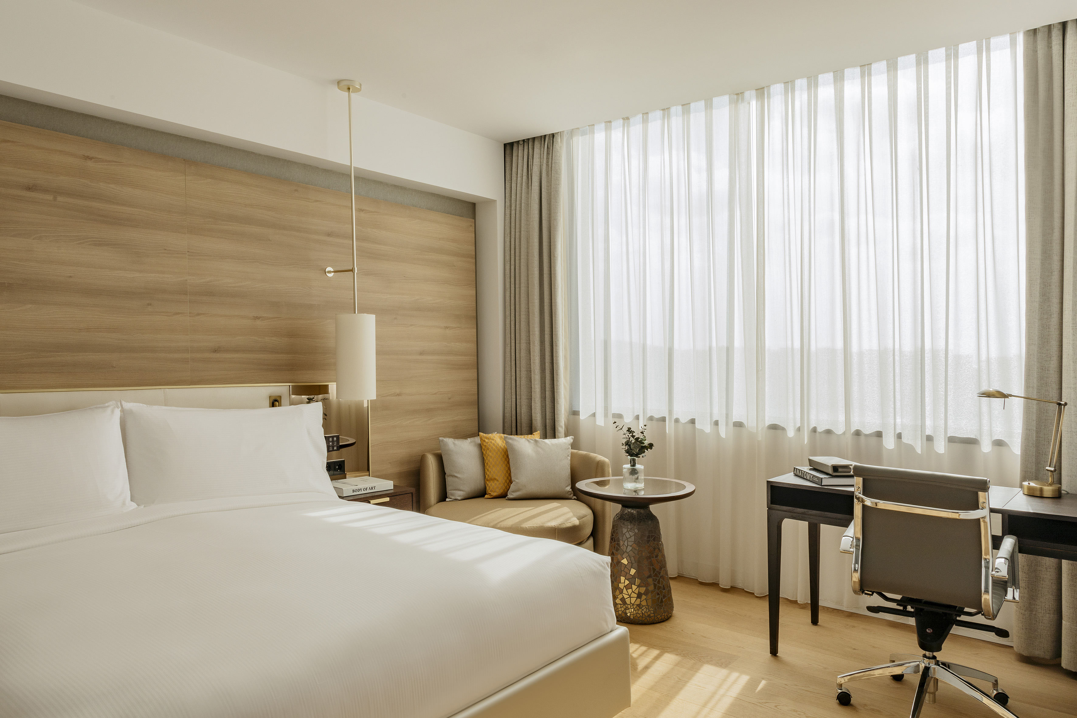 Stylish hotel room with white bed, wooden wall, desk, and bright window.