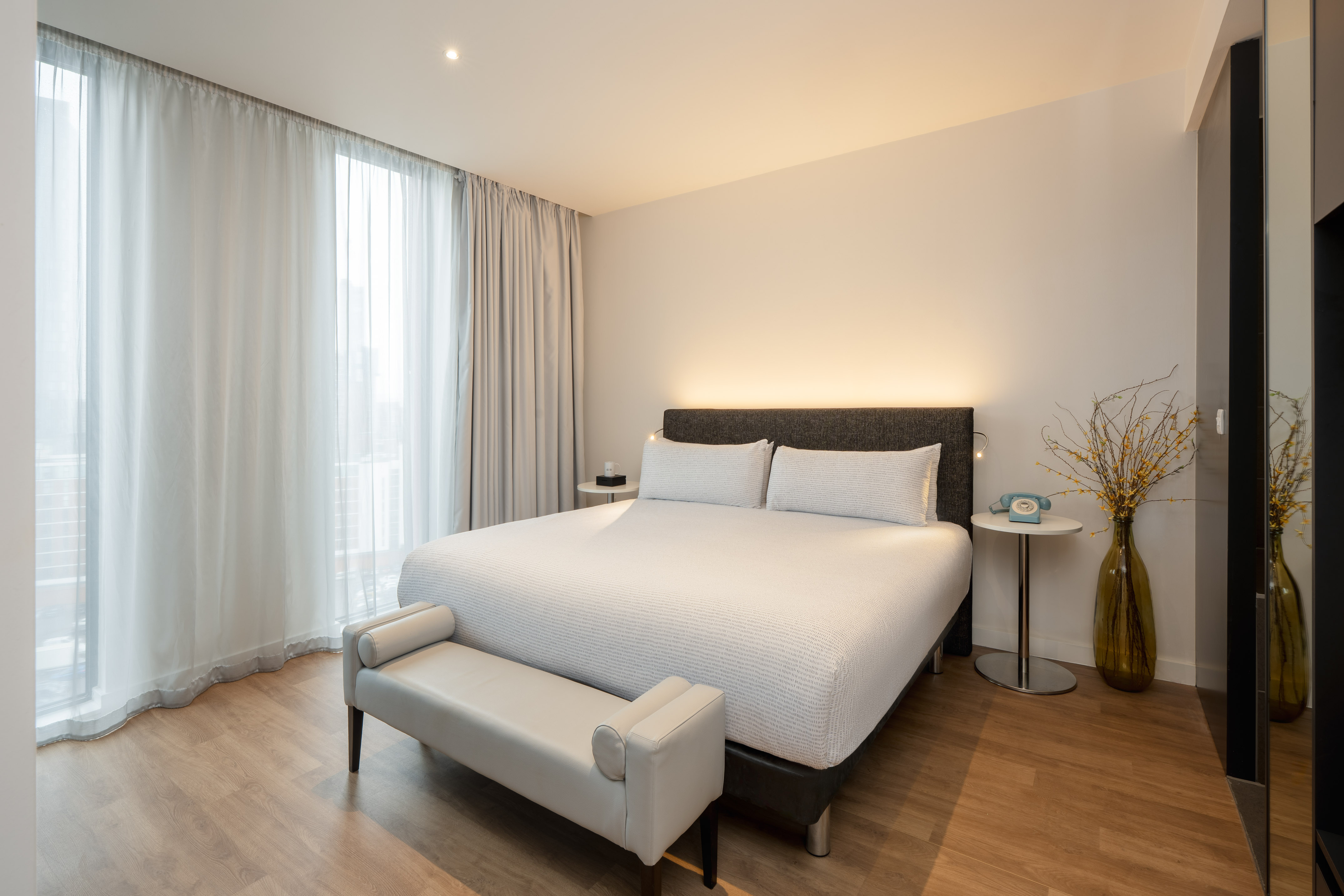 Modern hotel bedroom with king bed, white bedding, wood floor, and sheer curtains.