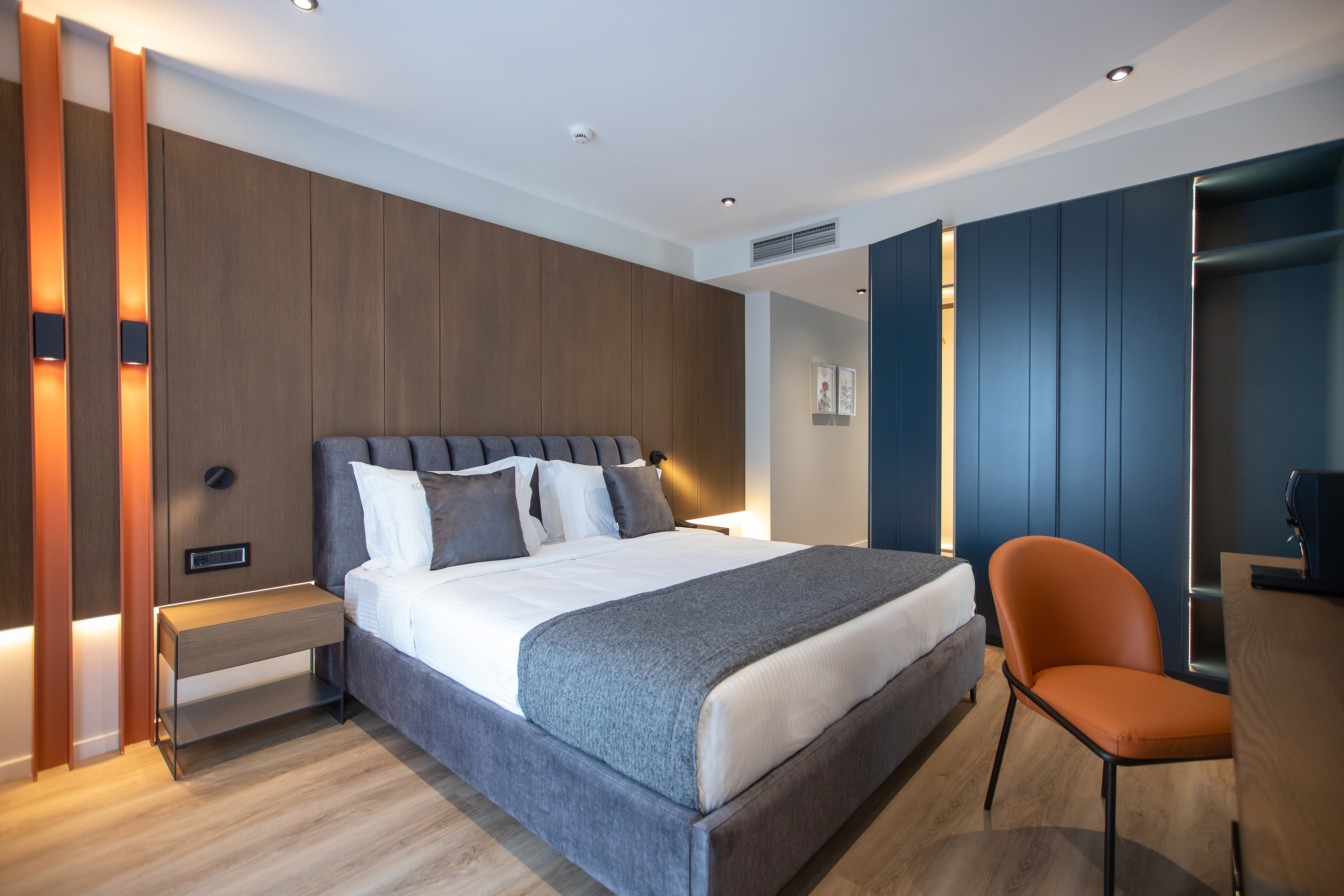 Contemporary hotel room: grey bed, wood wall, blue wardrobe, orange chair.