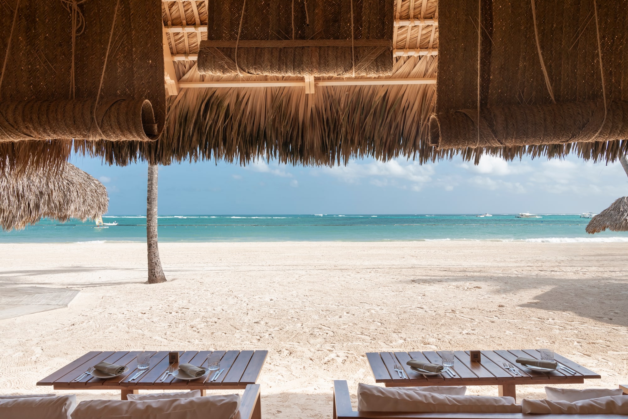 Restaurante de resort bajo palapa con mesas, frente a playa tropical.