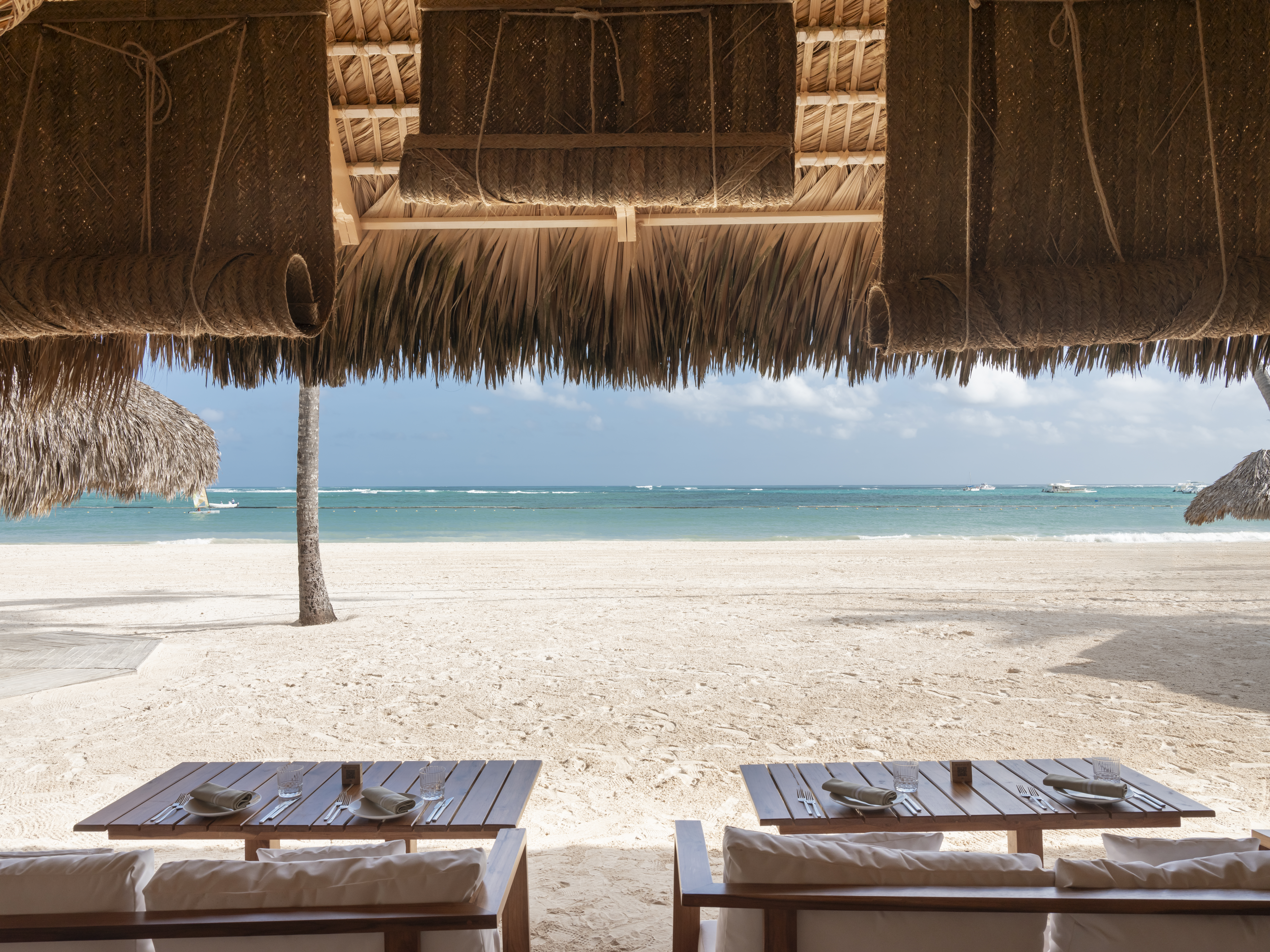 Restaurante de resort bajo palapa con mesas, frente a playa tropical.