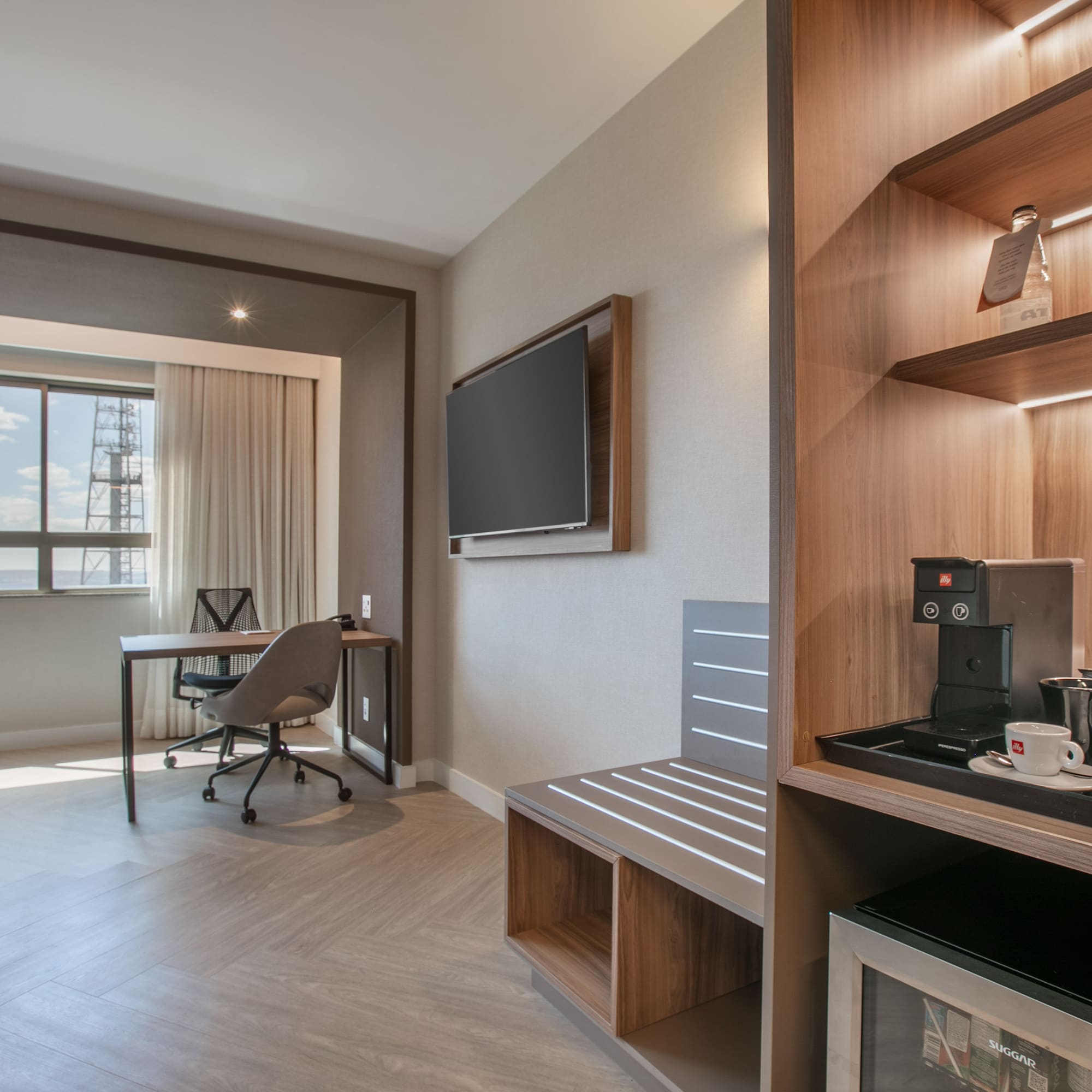 Modern hotel room with work desk, wall TV, coffee station, and large window.