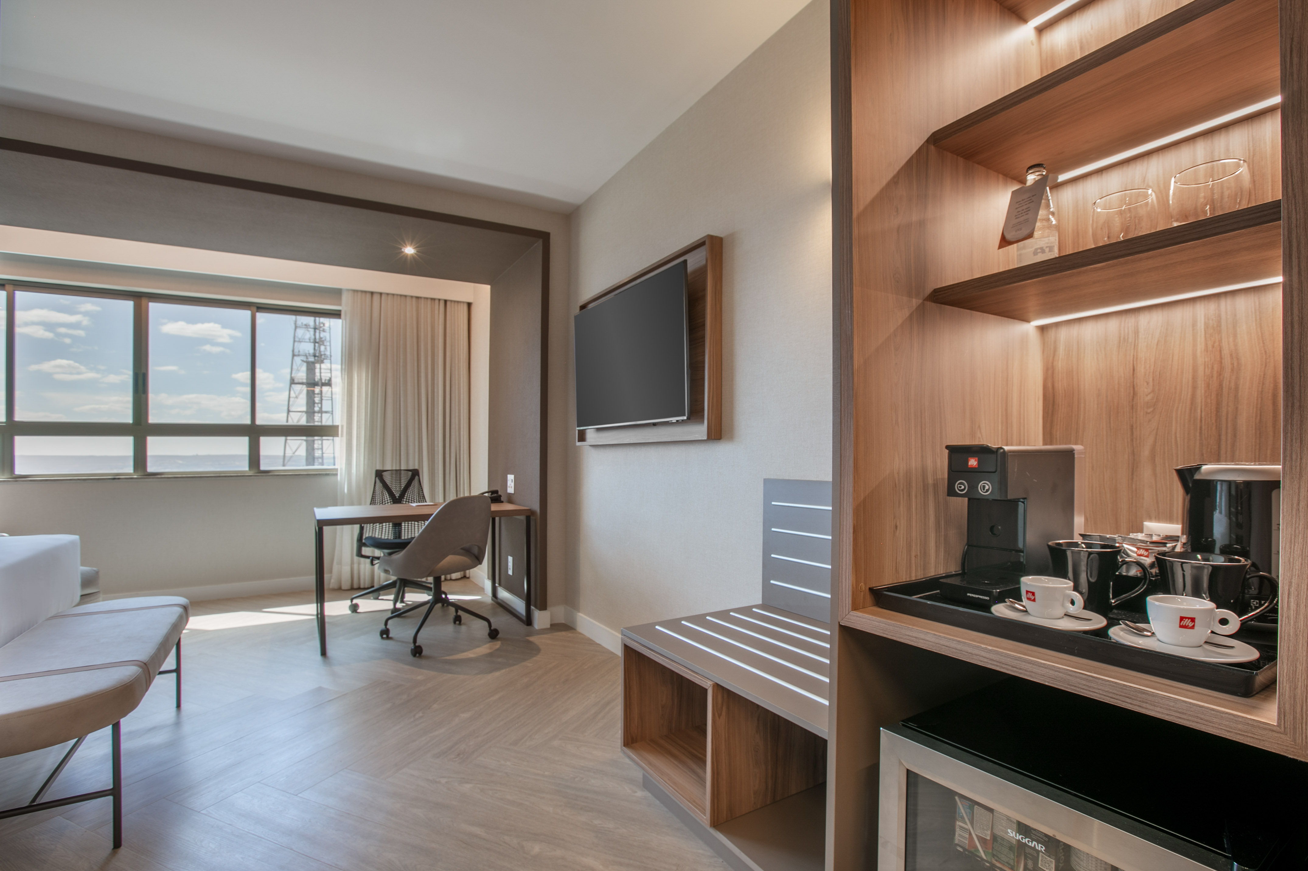 Modern hotel room with work desk, wall TV, coffee station, and large window.