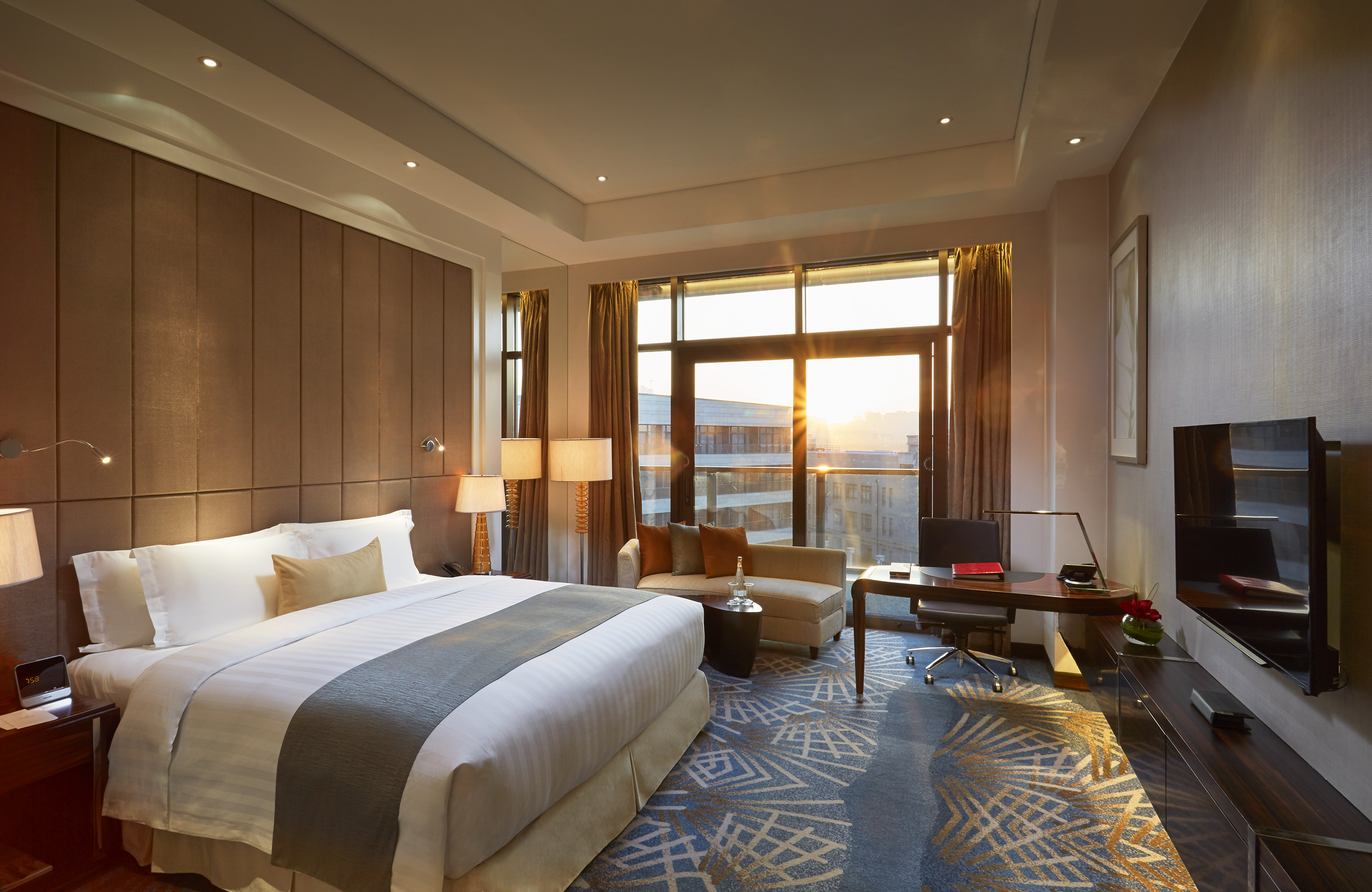 Luxurious hotel room with king bed, desk, sofa, and sunlit city view.
