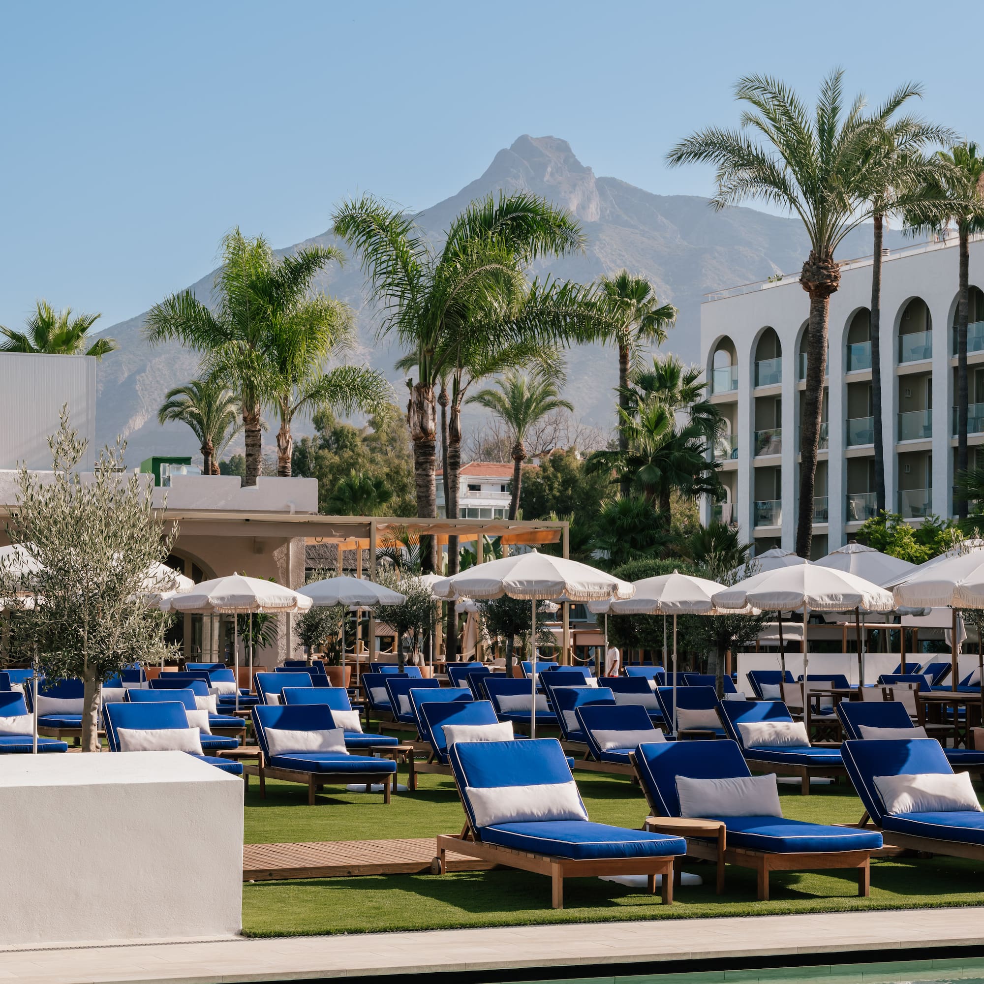Marbella resort pool area with blue sun loungers, white umbrellas, and mountain view.