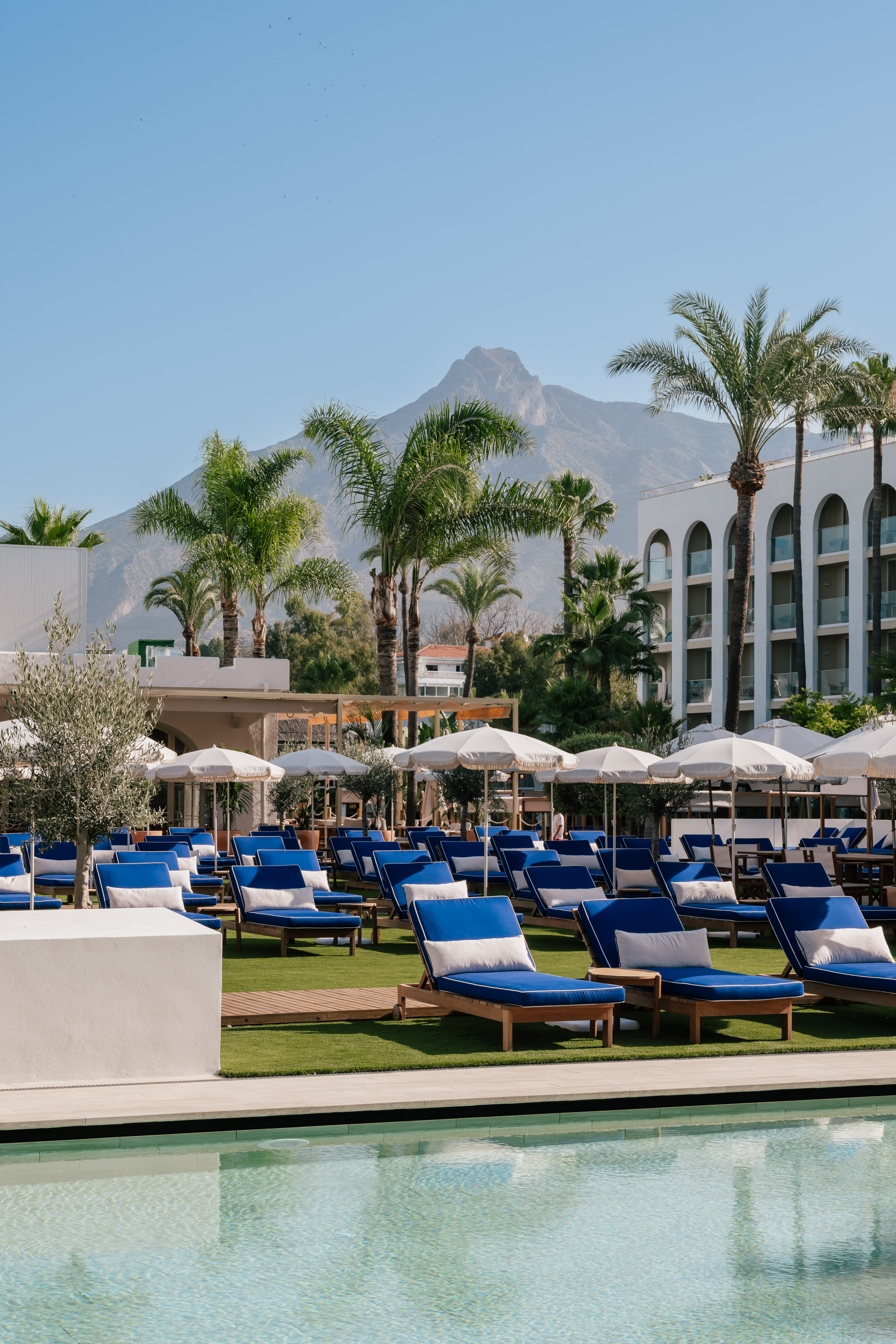 Marbella resort pool area with blue sun loungers, white umbrellas, and mountain view.