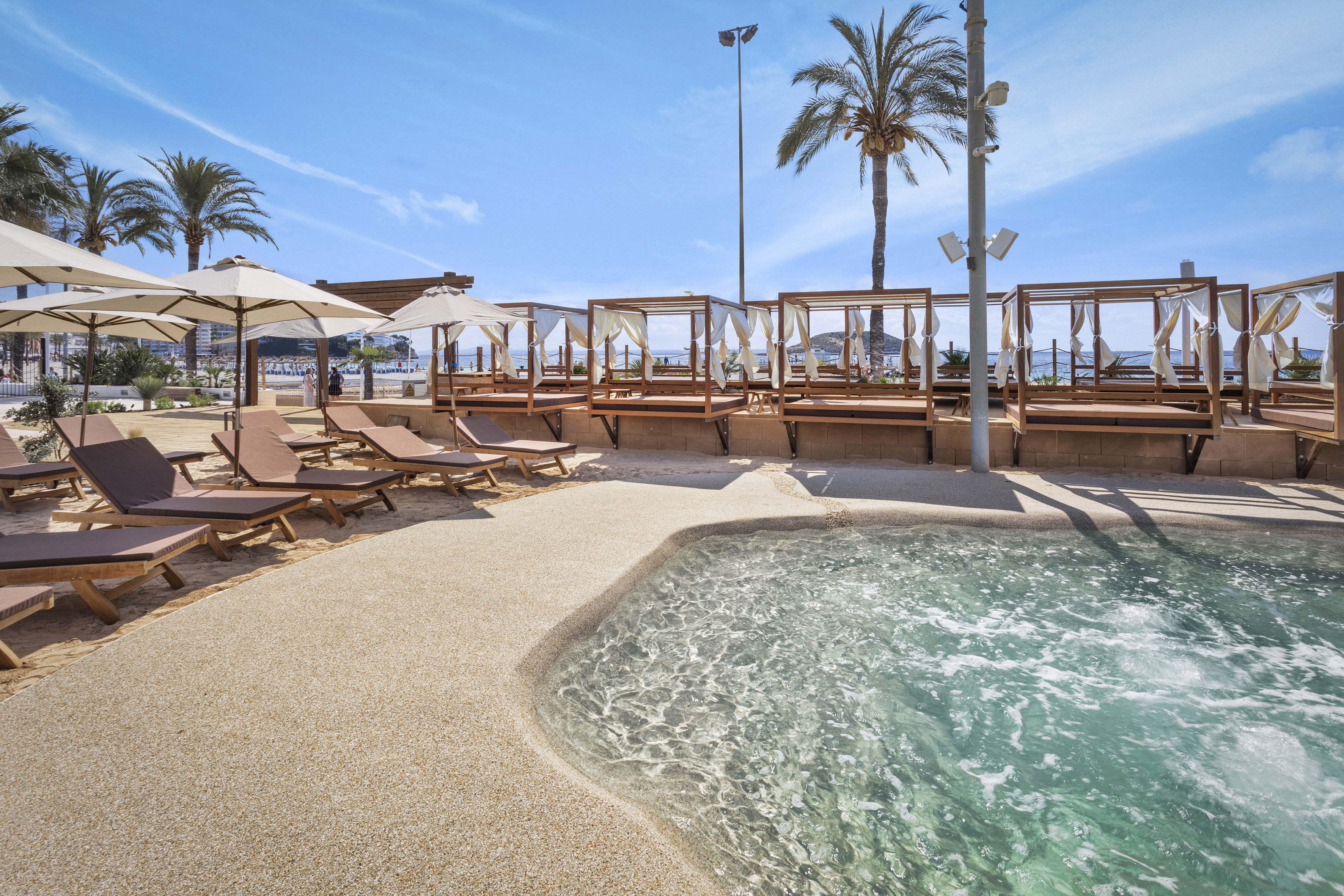 Sunny beach club with clear pool, cabanas, sun loungers, and palm trees.