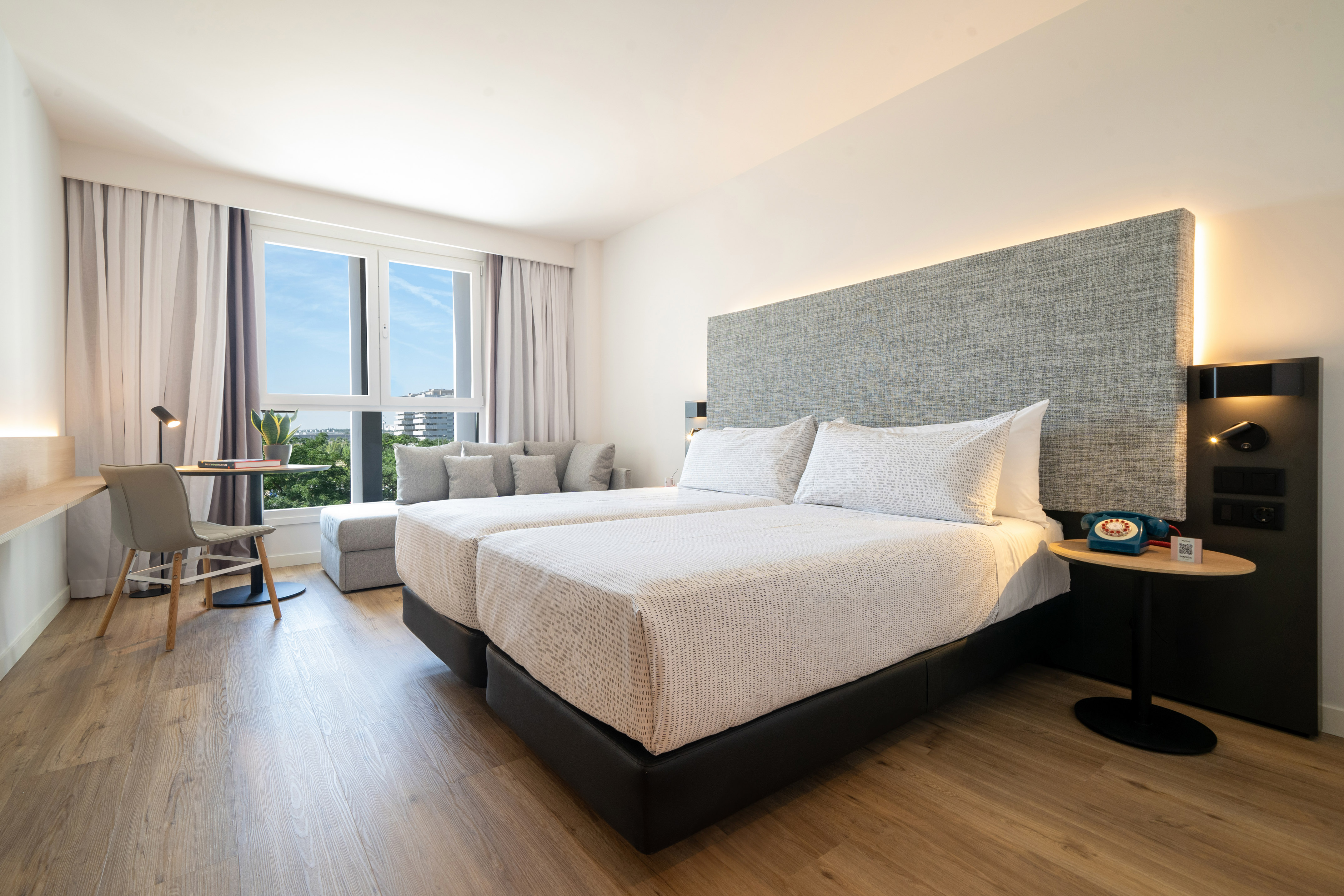 Modern hotel room with twin beds, bright window, sofa, and city view.