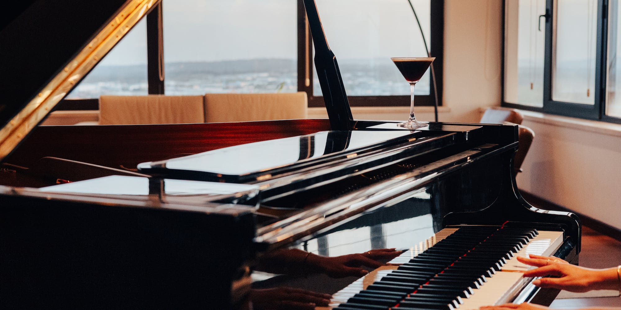 Hands playing a polished grand piano, cocktail on top, cityscape background.