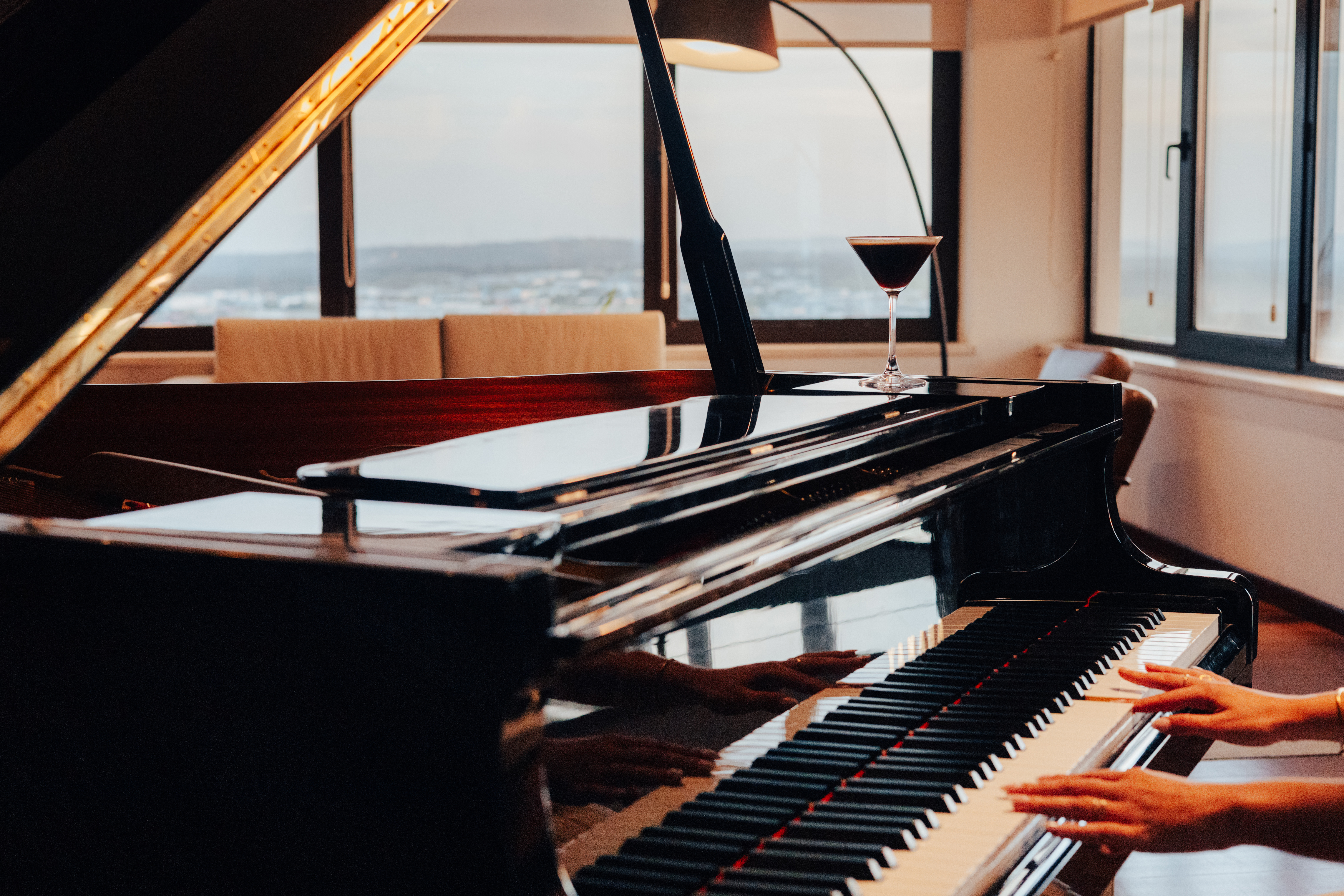Hands playing a polished grand piano, cocktail on top, cityscape background.
