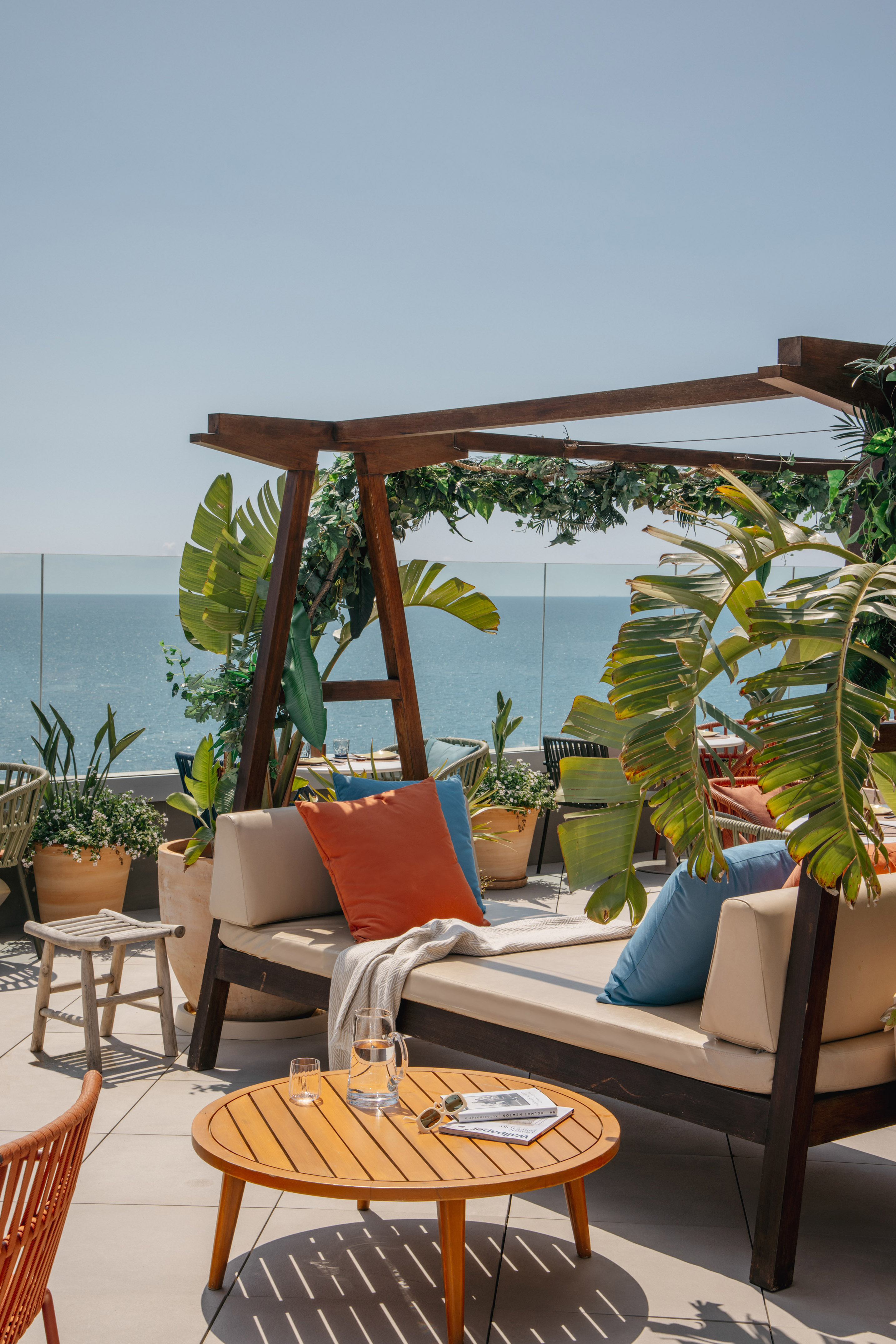 Oceanfront rooftop lounge with daybed, tropical plants, and wooden table.