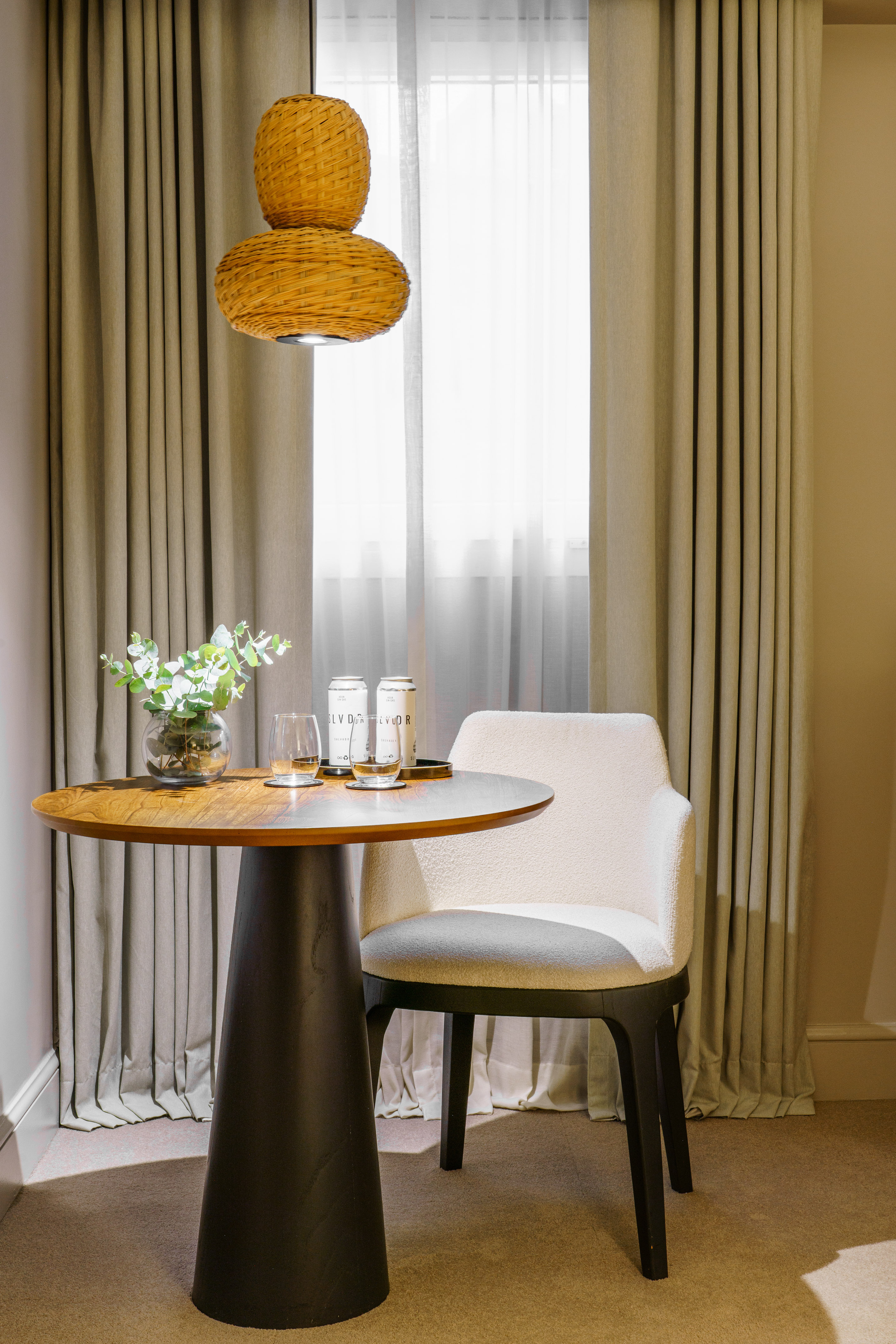 Modern hotel room corner with wooden table, cream chair, drinks, woven light.