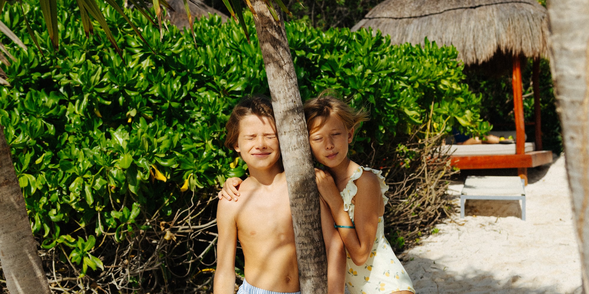 Children on tropical beach leaning on palm tree, sandy shore, resort hut background.