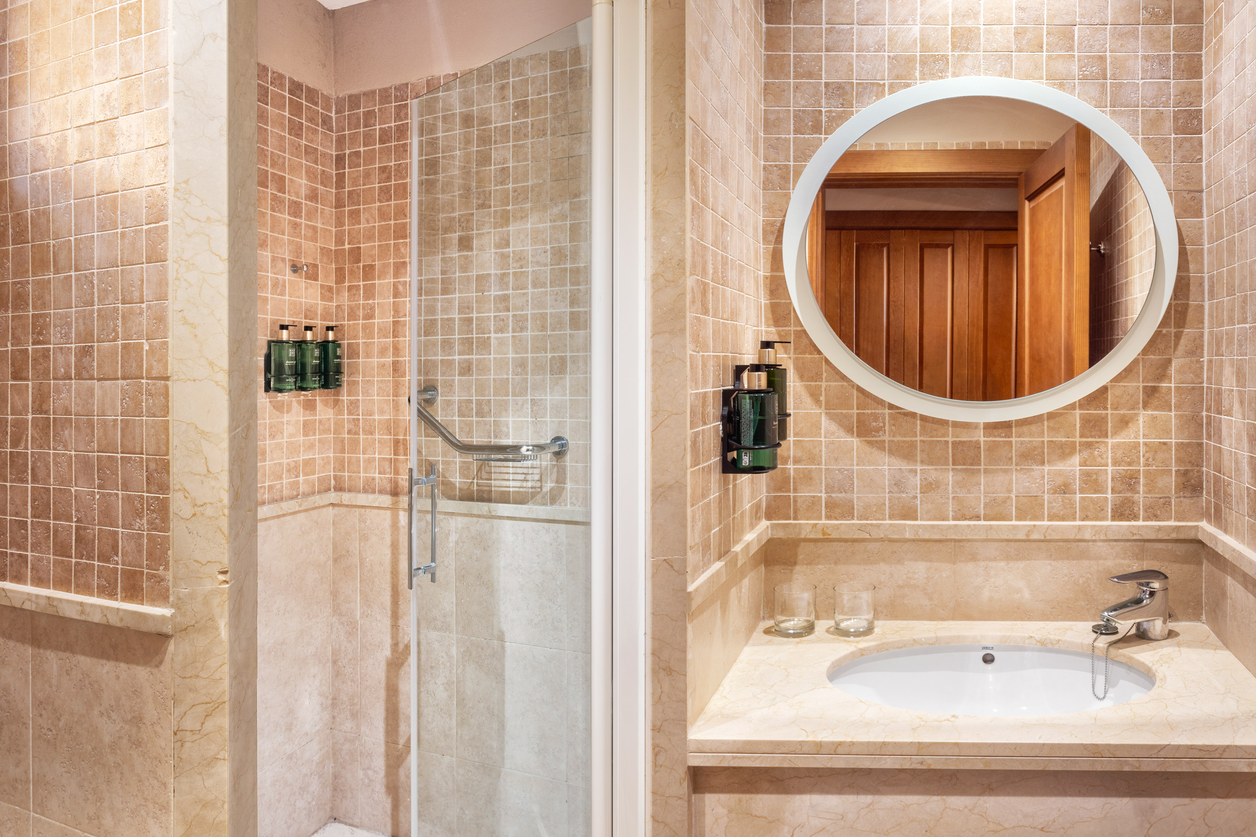 Beige tiled bathroom with shower stall, sink vanity, and round mirror.