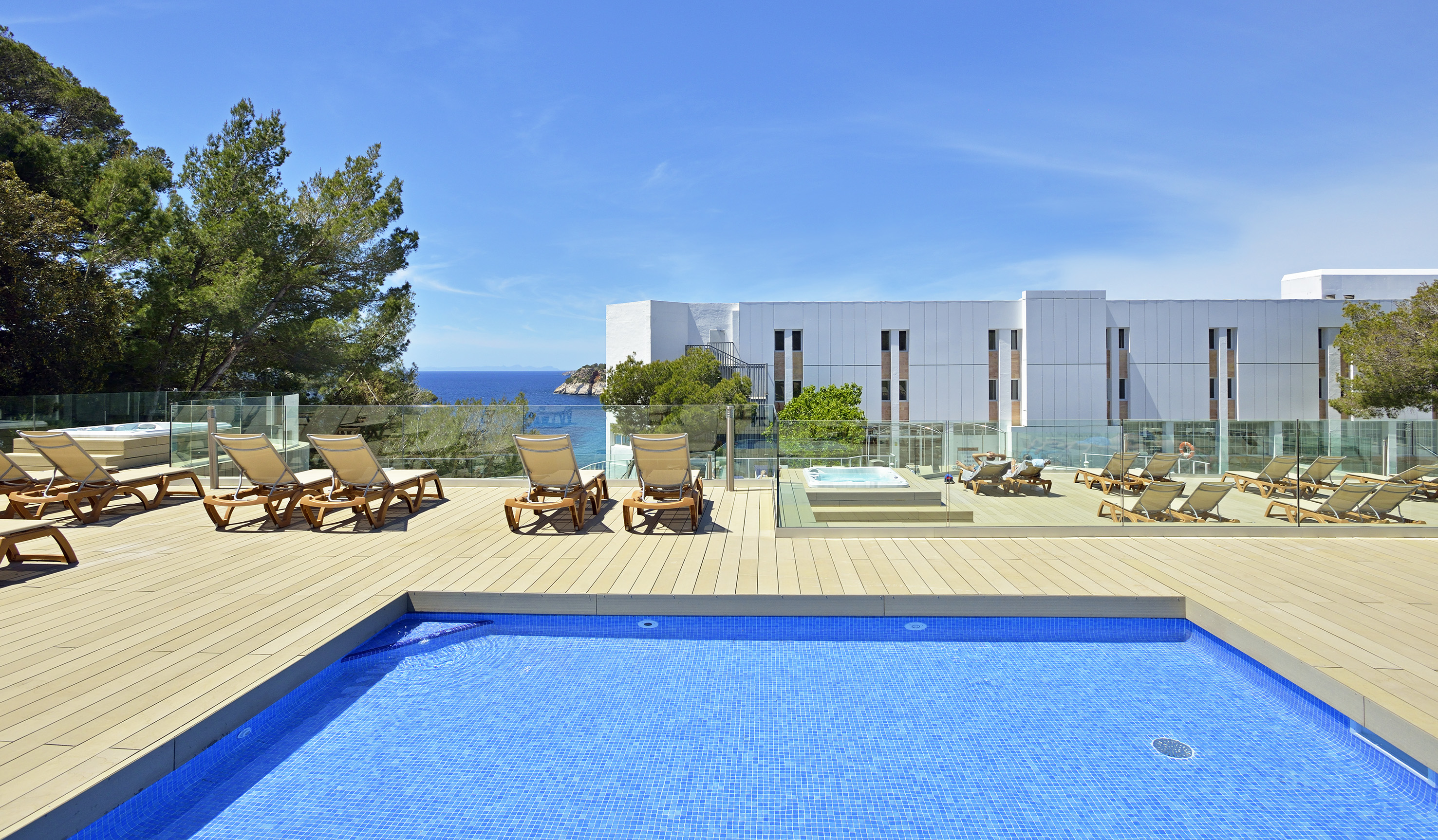 Sunny hotel pool deck with ocean view, sun loungers, and hot tub.