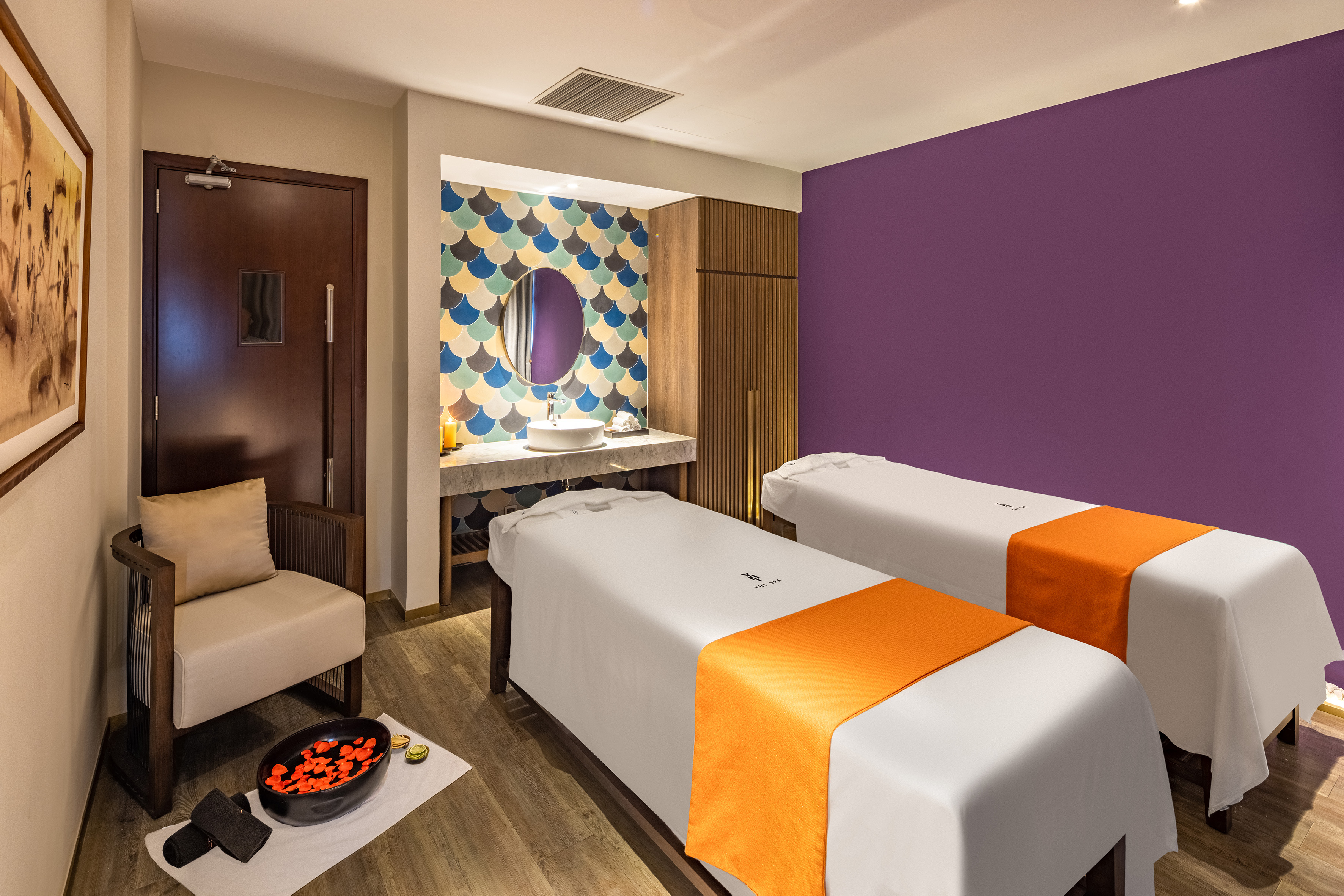 Modern spa room with two massage beds, purple wall, and stylish tiled sink area.