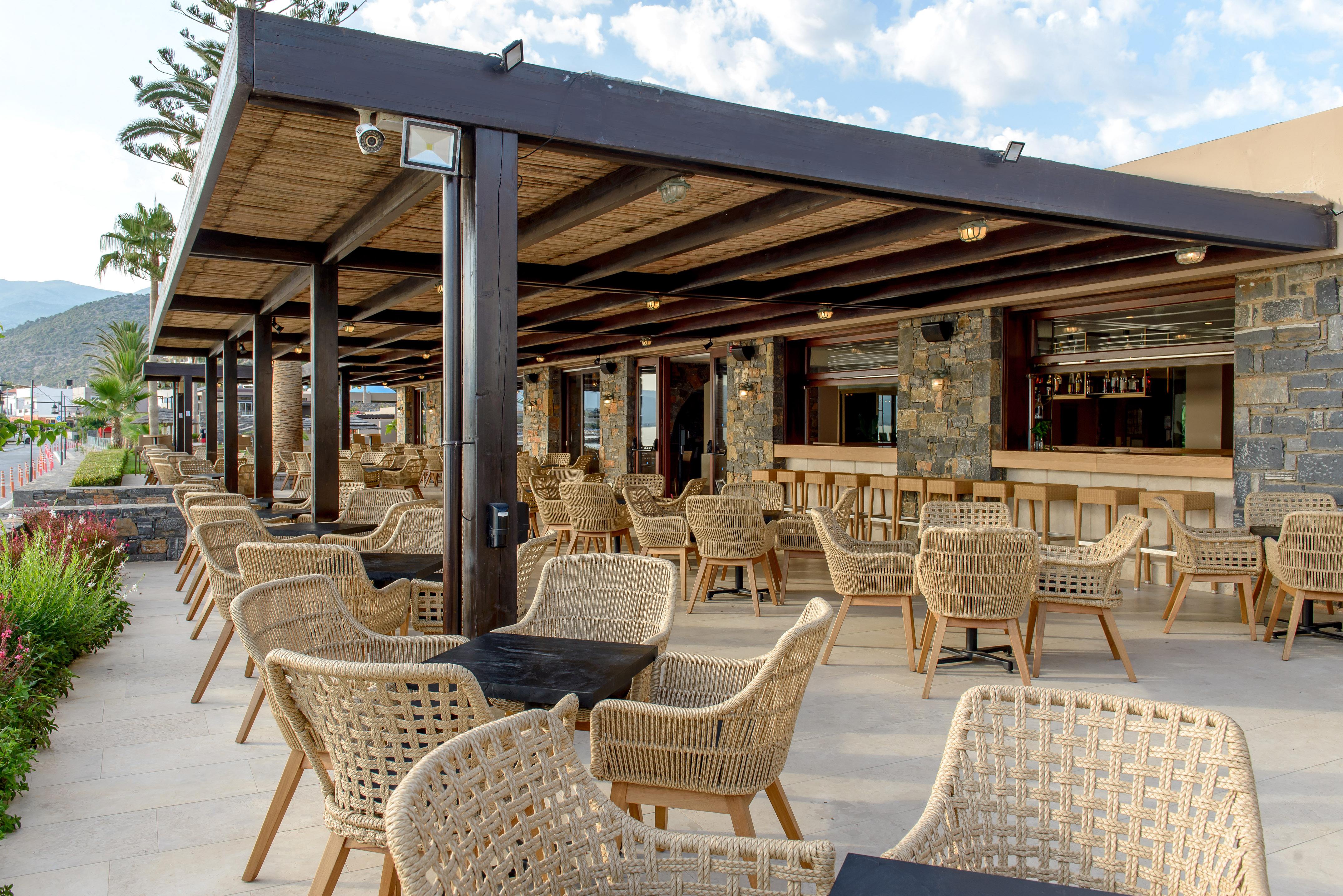 Outdoor restaurant terrace with woven chairs, dark tables, and stone bar area.