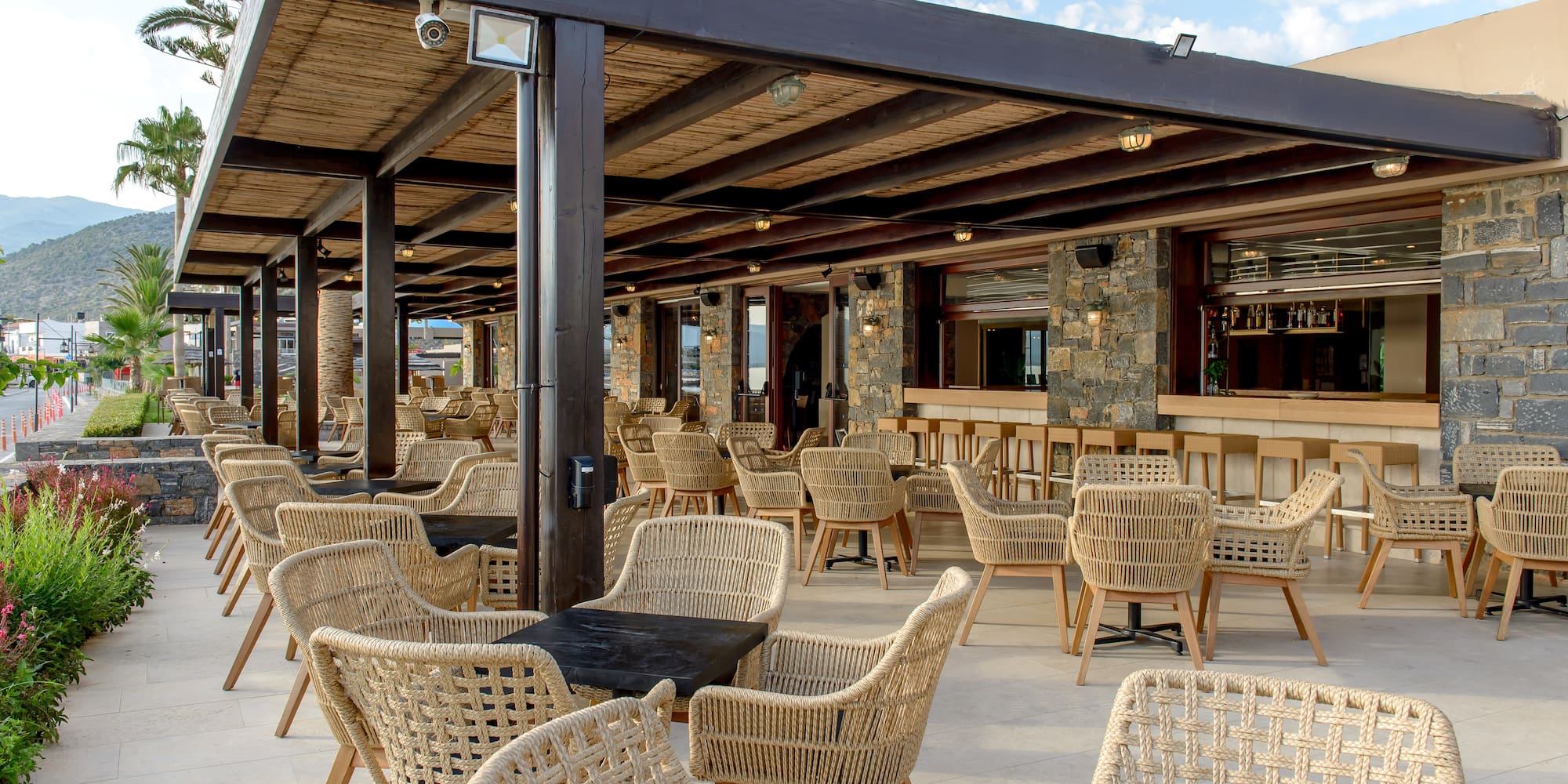 Outdoor restaurant terrace with woven chairs, dark tables, and stone bar area.