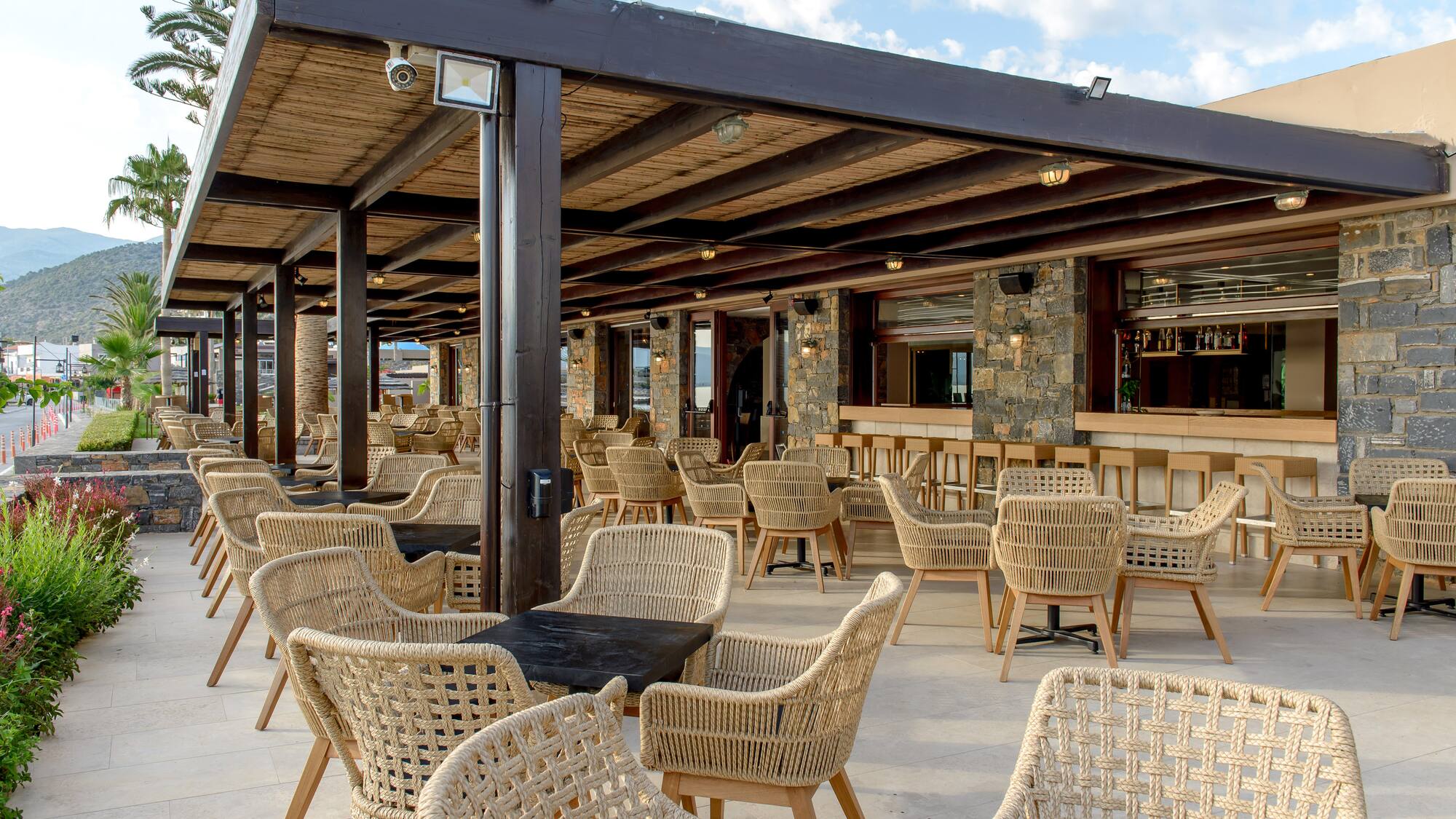 Outdoor restaurant terrace with woven chairs, dark tables, and stone bar area.