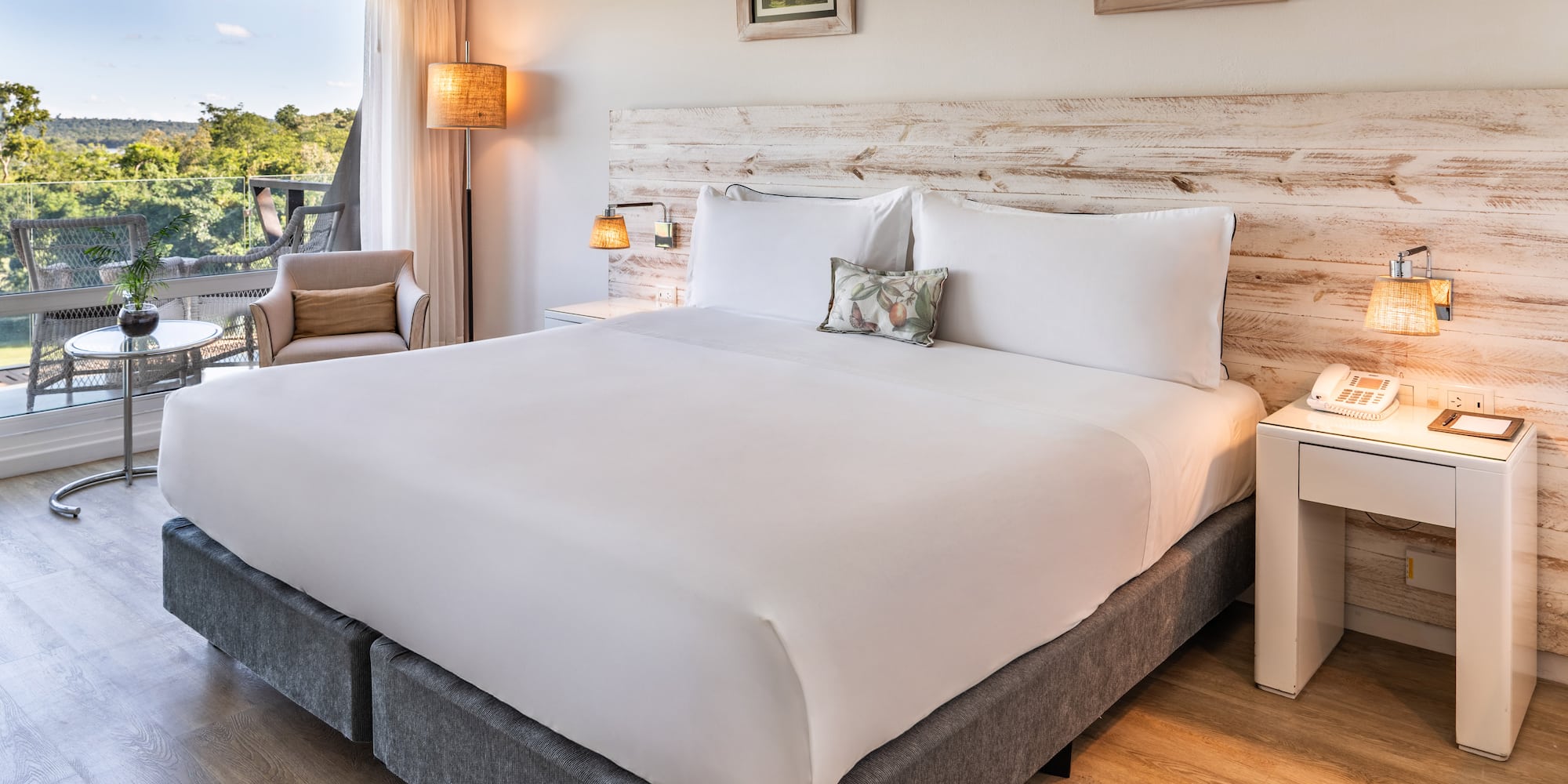 a bed with white sheets and pillows in a room with a deck and chairs