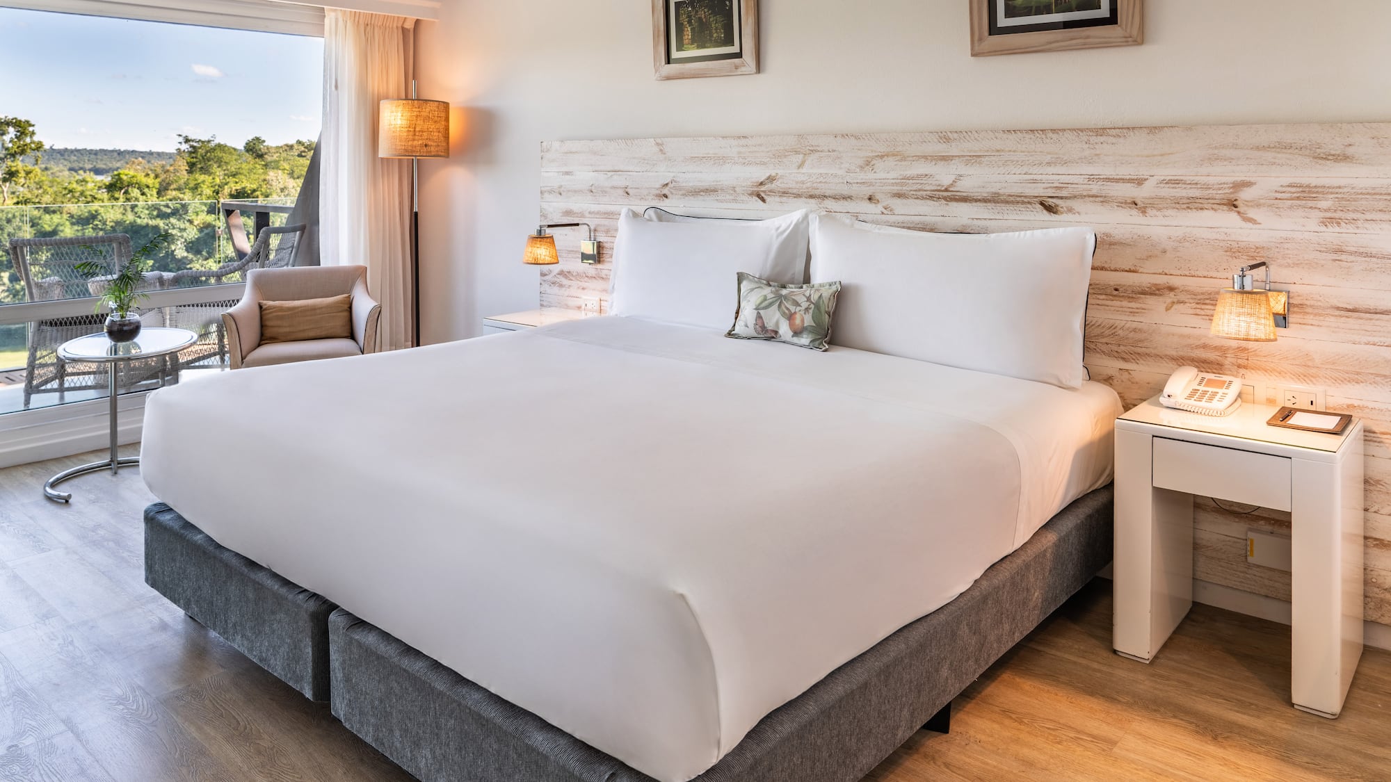 a bed with white sheets and pillows in a room with a deck and chairs