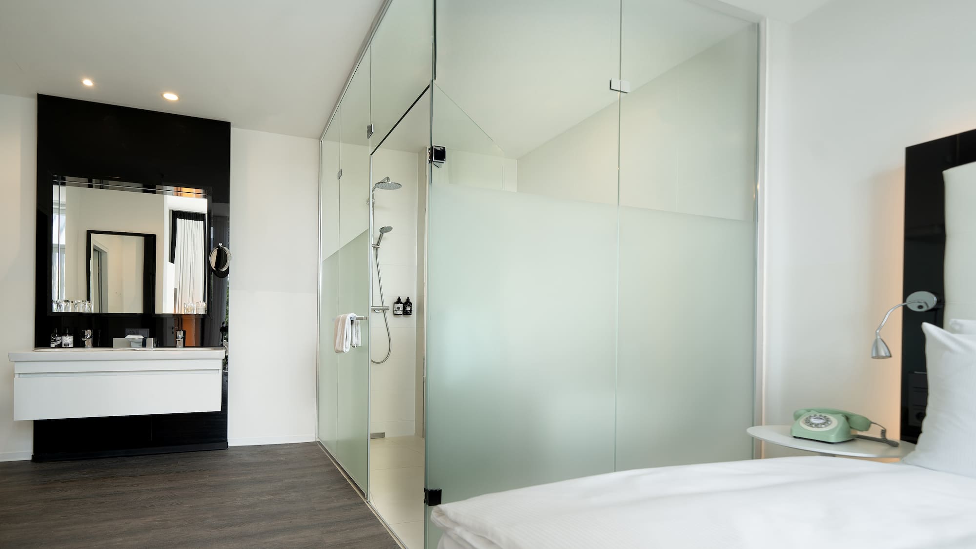 Contemporary hotel room with glass shower, vanity, and white bed.