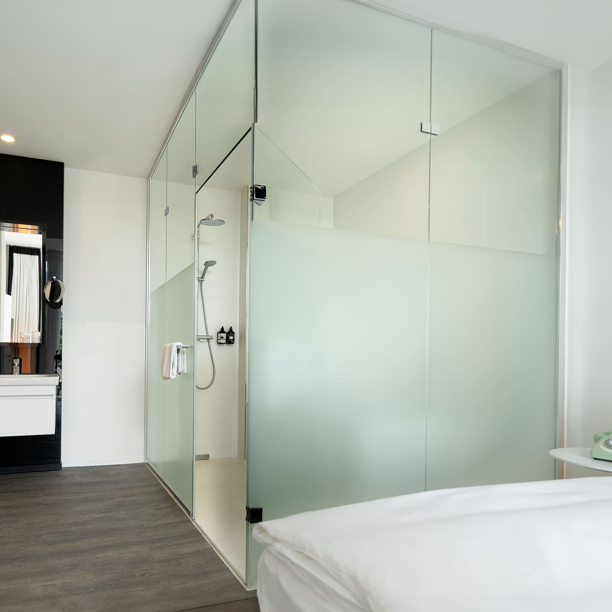 Contemporary hotel room with glass shower, vanity, and white bed.