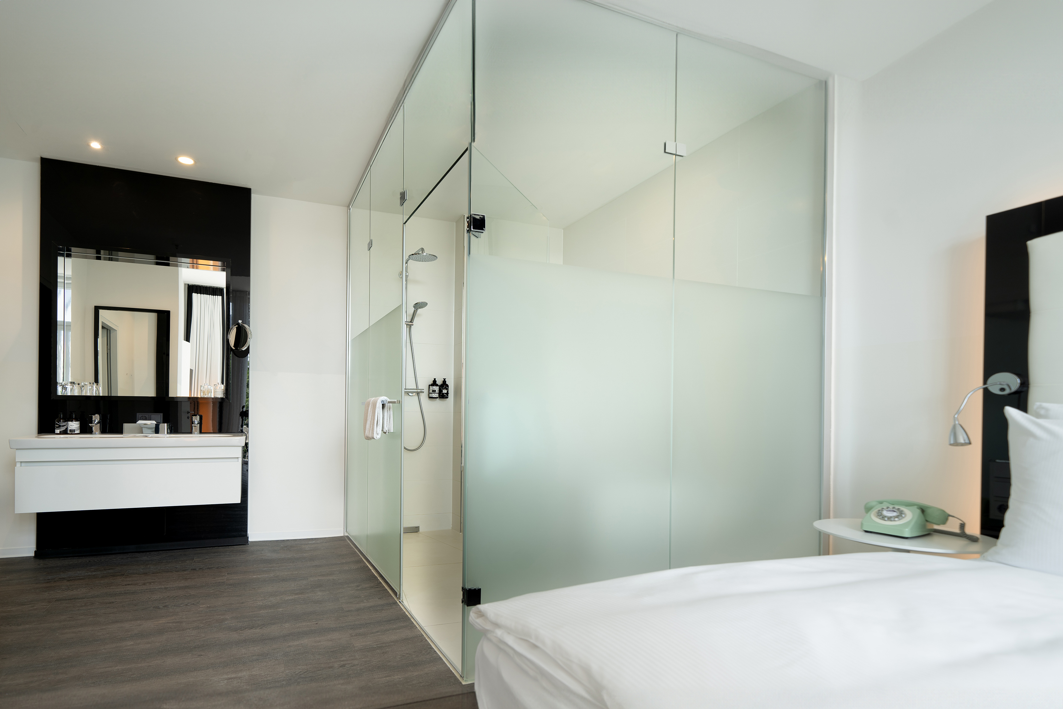 Contemporary hotel room with glass shower, vanity, and white bed.