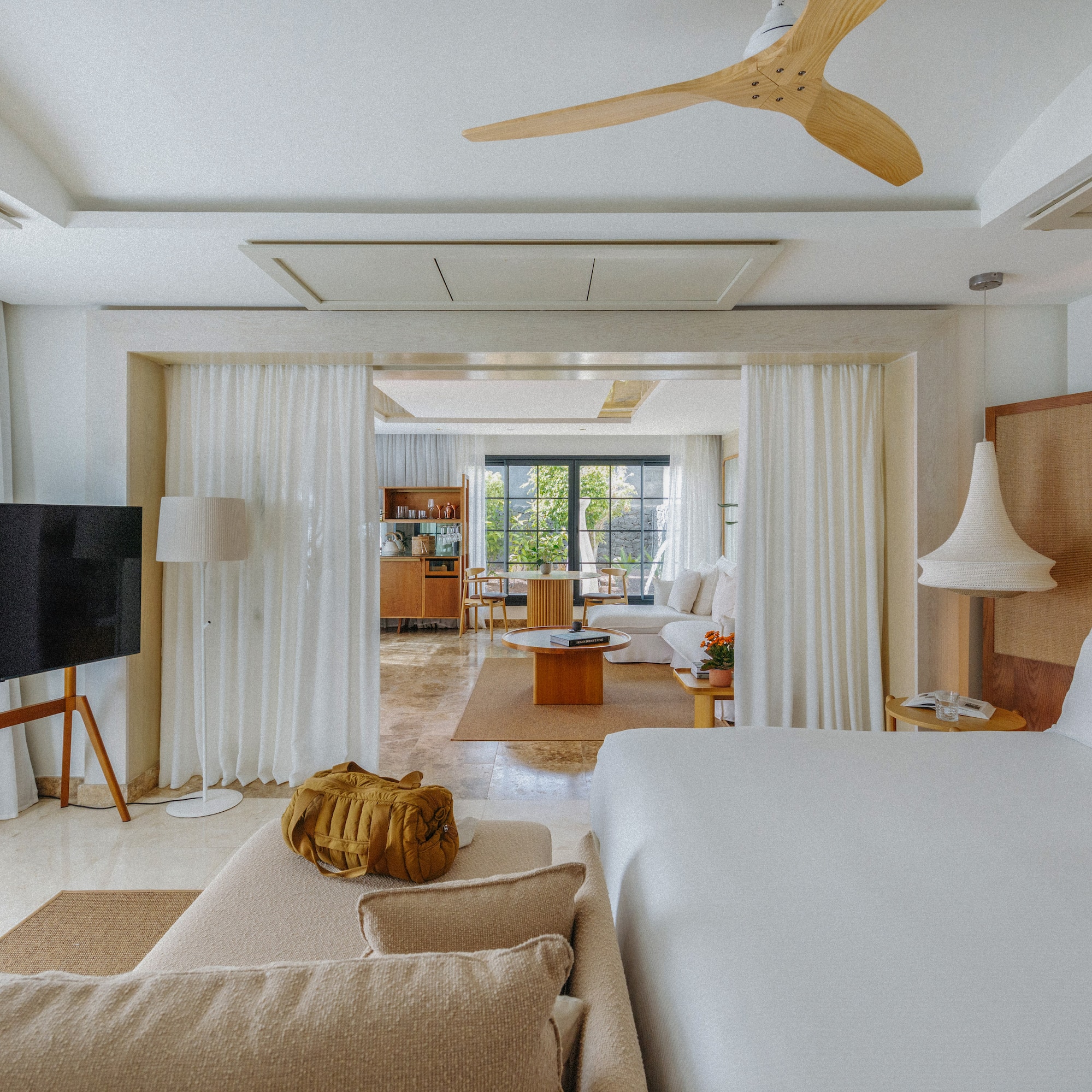 Open-concept hotel suite with bedroom, living area, white decor, and wooden fan.