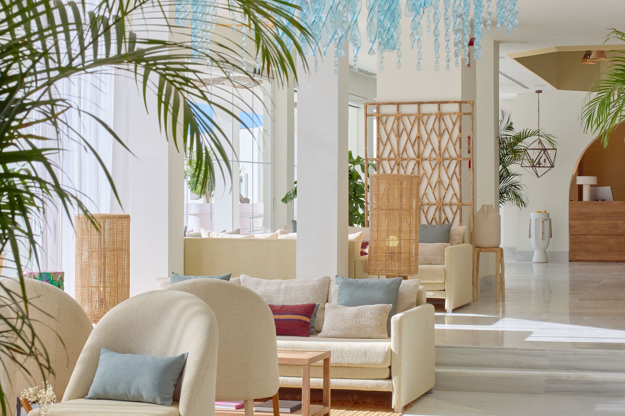 Elegant hotel lounge with light furniture, palm plants, and blue ceiling art.