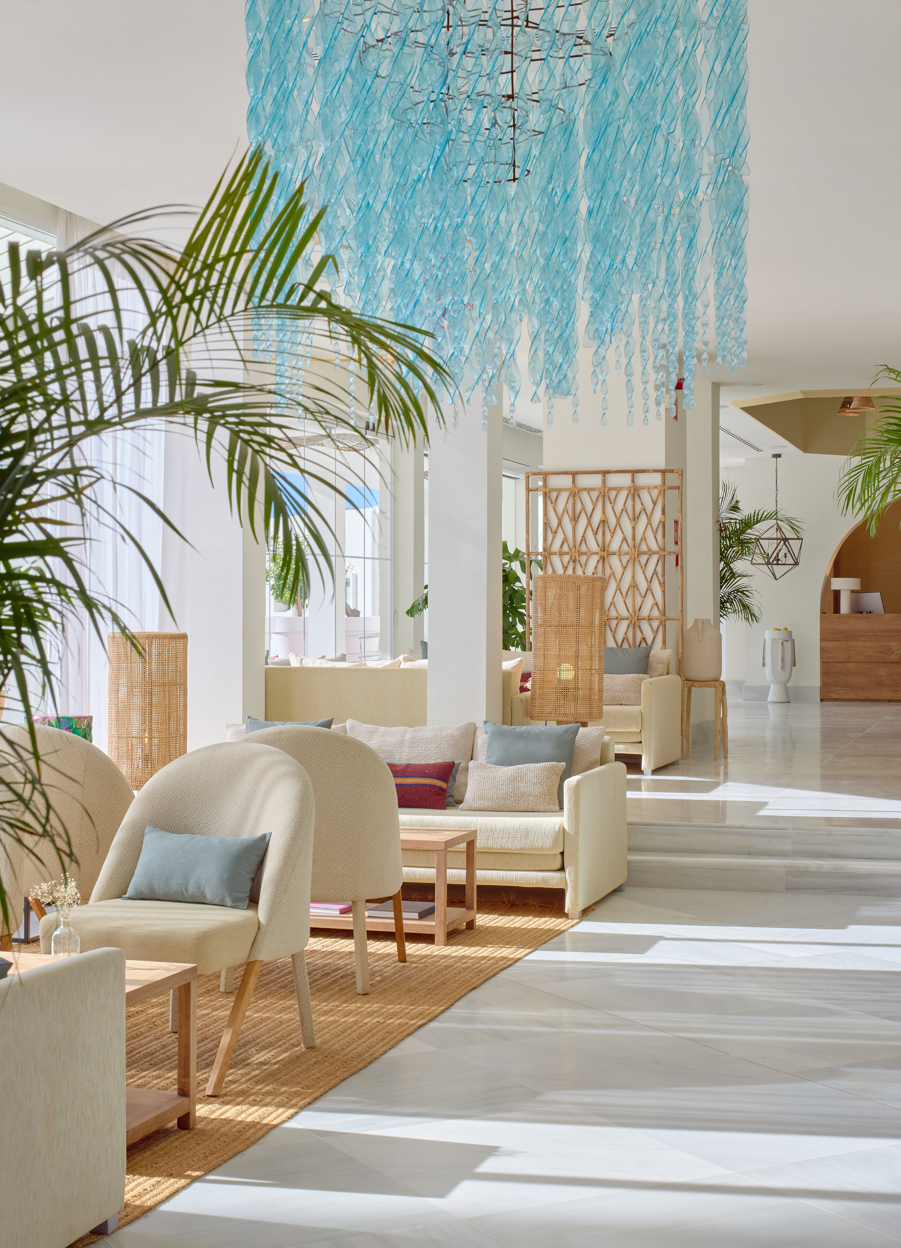Elegant hotel lounge with light furniture, palm plants, and blue ceiling art.
